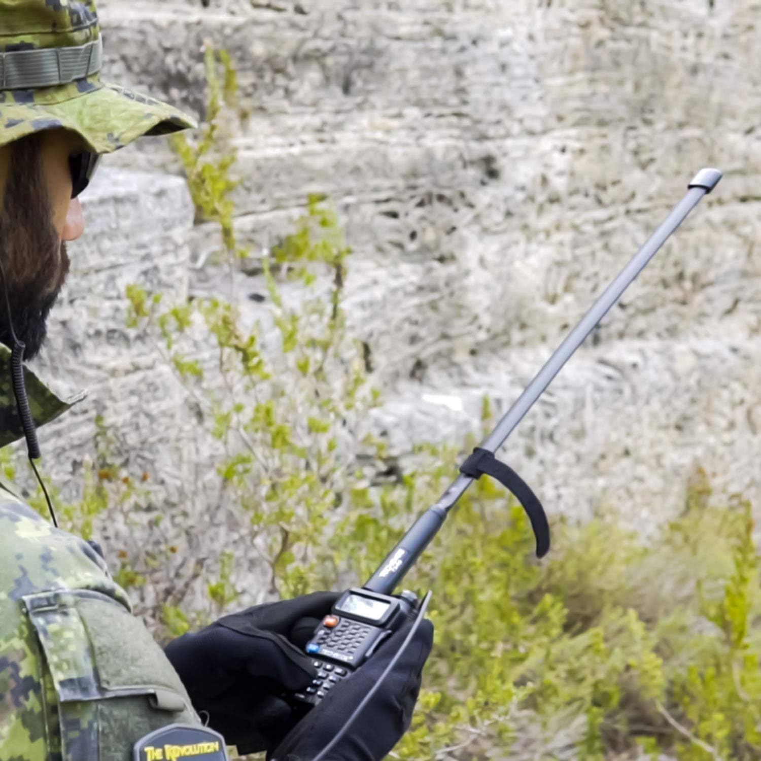 Soldato in mimetica che tiene una radio con antenna, vicino a rocce e cespugli verdi.
