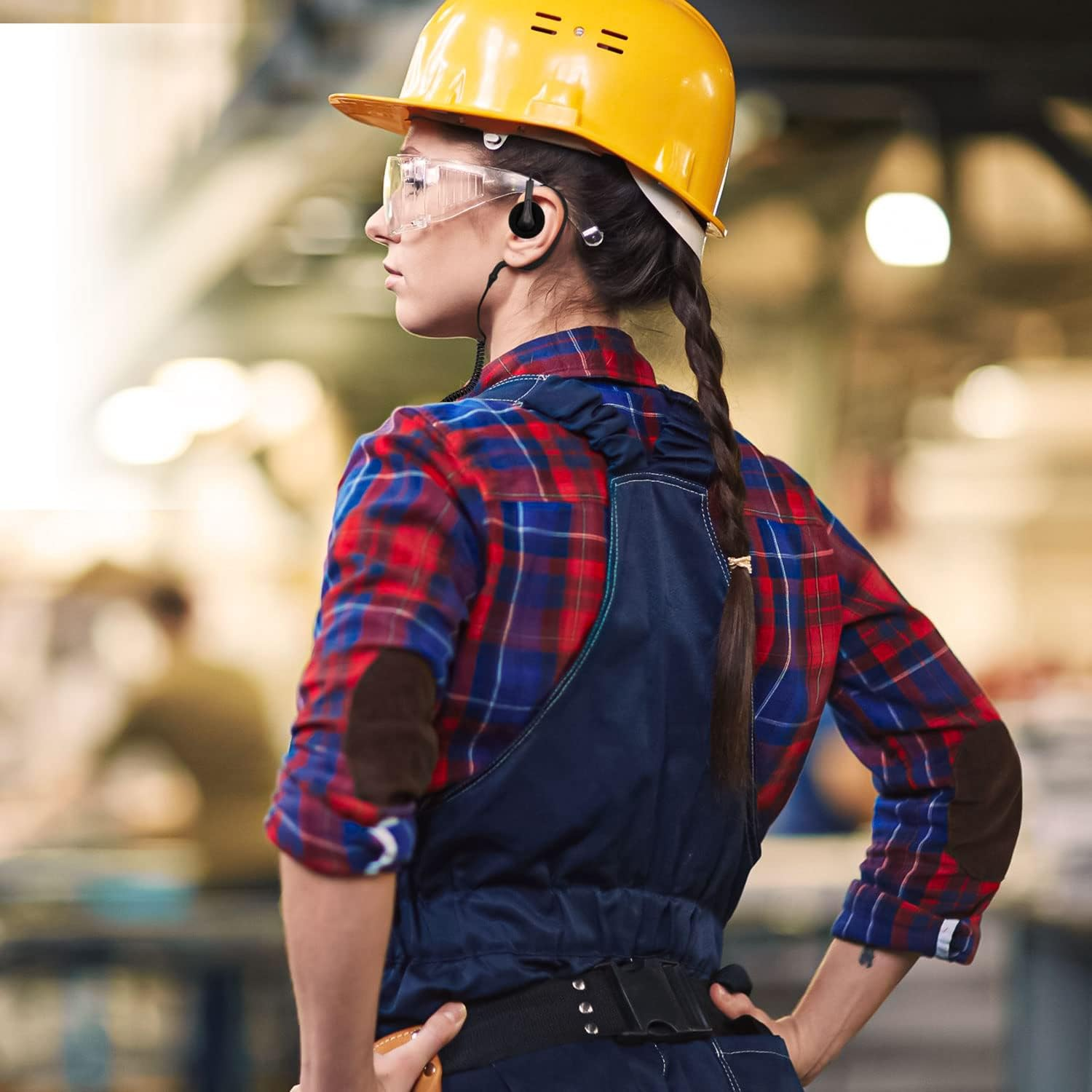 Una donna con casco giallo, occhiali protettivi e camicia a quadri è in una fabbrica. Ha le mani sui fianchi.