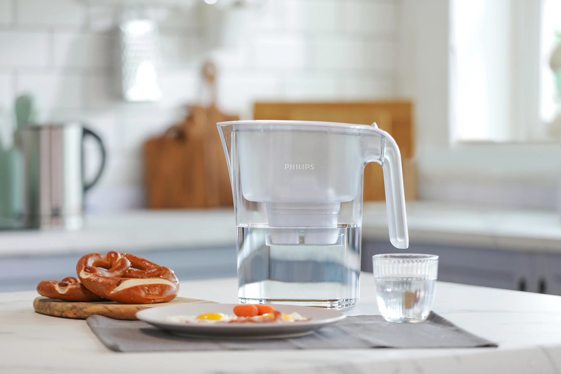 Una caraffa filtrante Philips con acqua, un bicchiere d'acqua, pretzel e uova fritte su un tavolo in cucina.