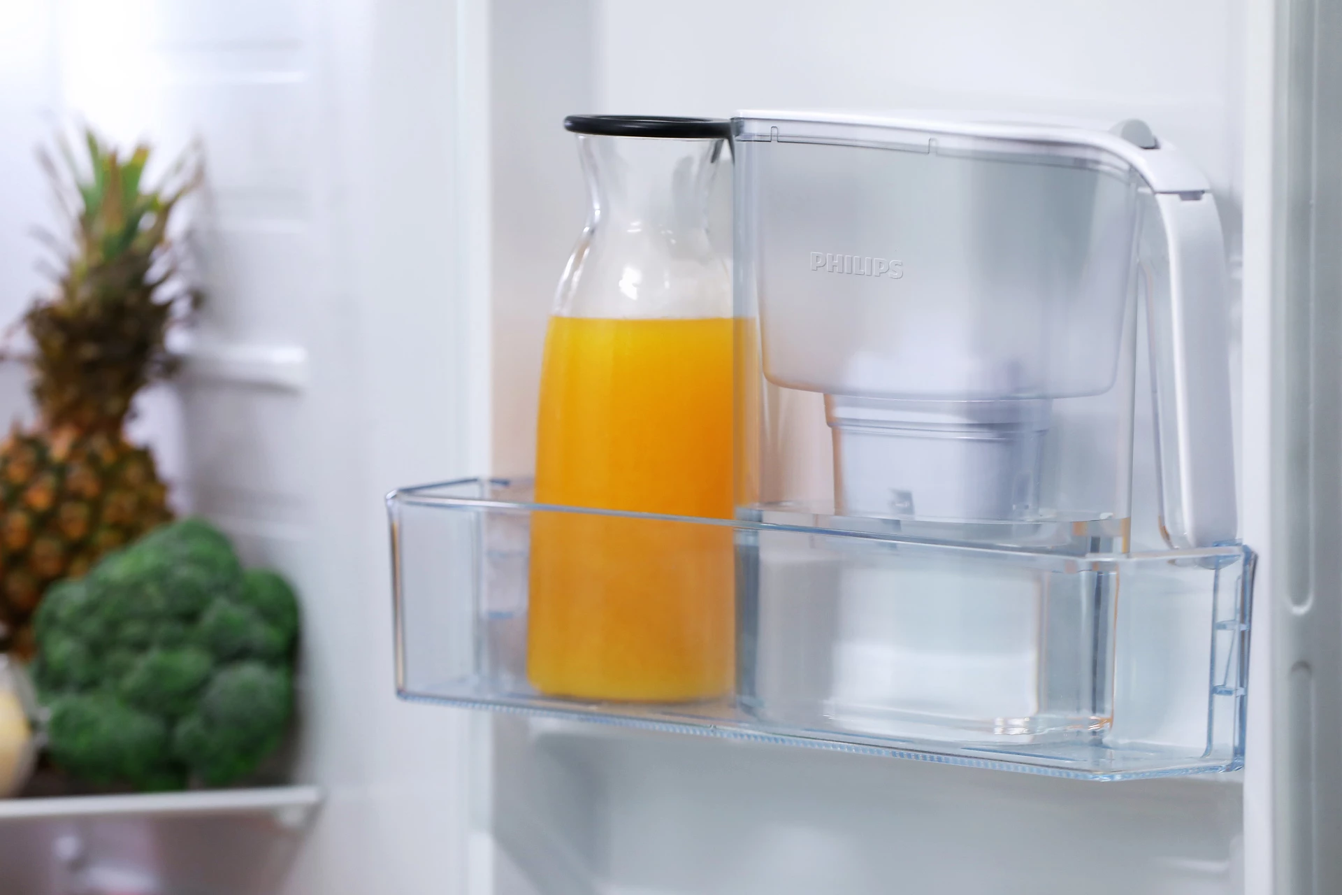 Un ripiano del frigorifero con una caraffa di succo d'arancia e un filtro dell'acqua Philips. Frutta come ananas e broccoli sono visibili.