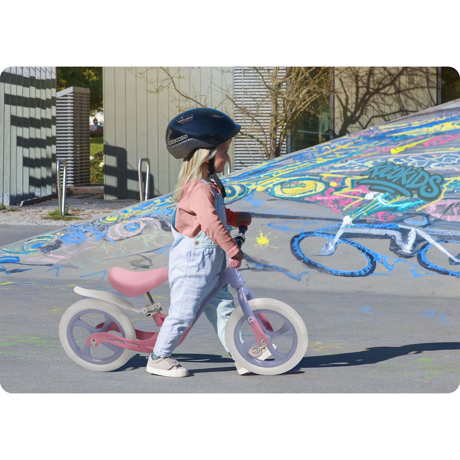 Mała dziewczynka w kasku jeździ na różowym rowerku biegowym po szarej powierzchni. Graffiti w tle.