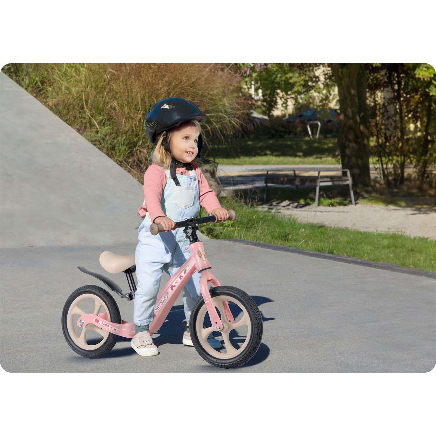Mała dziewczynka w kasku jeździ na różowym rowerku biegowym po betonie. Za nią zieleń i ławka w parku.
