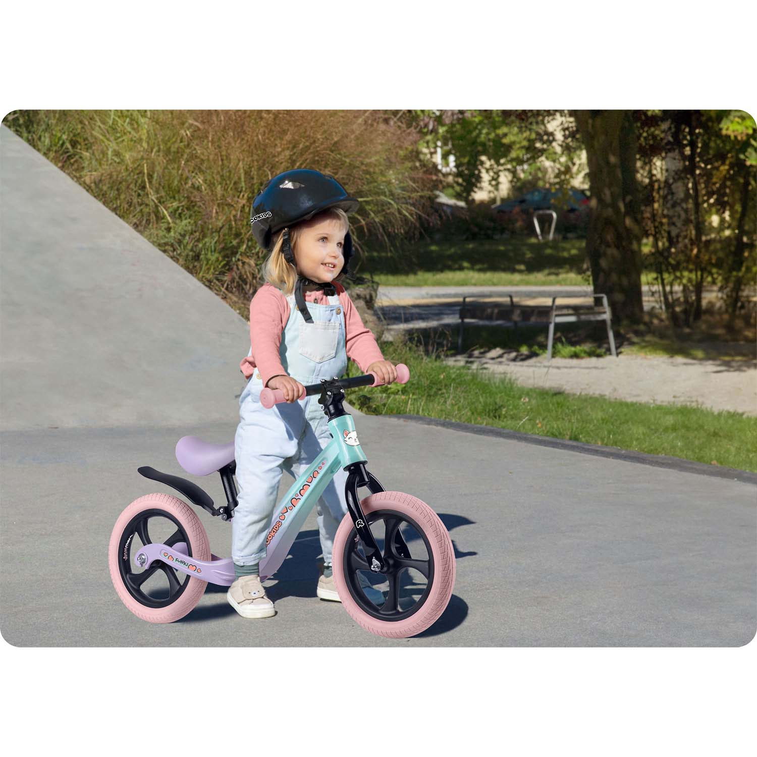 Mała dziewczynka w kasku jeździ na rowerze biegowym po utwardzonej nawierzchni. Rower jest jasnoniebieski z różowymi kołami.