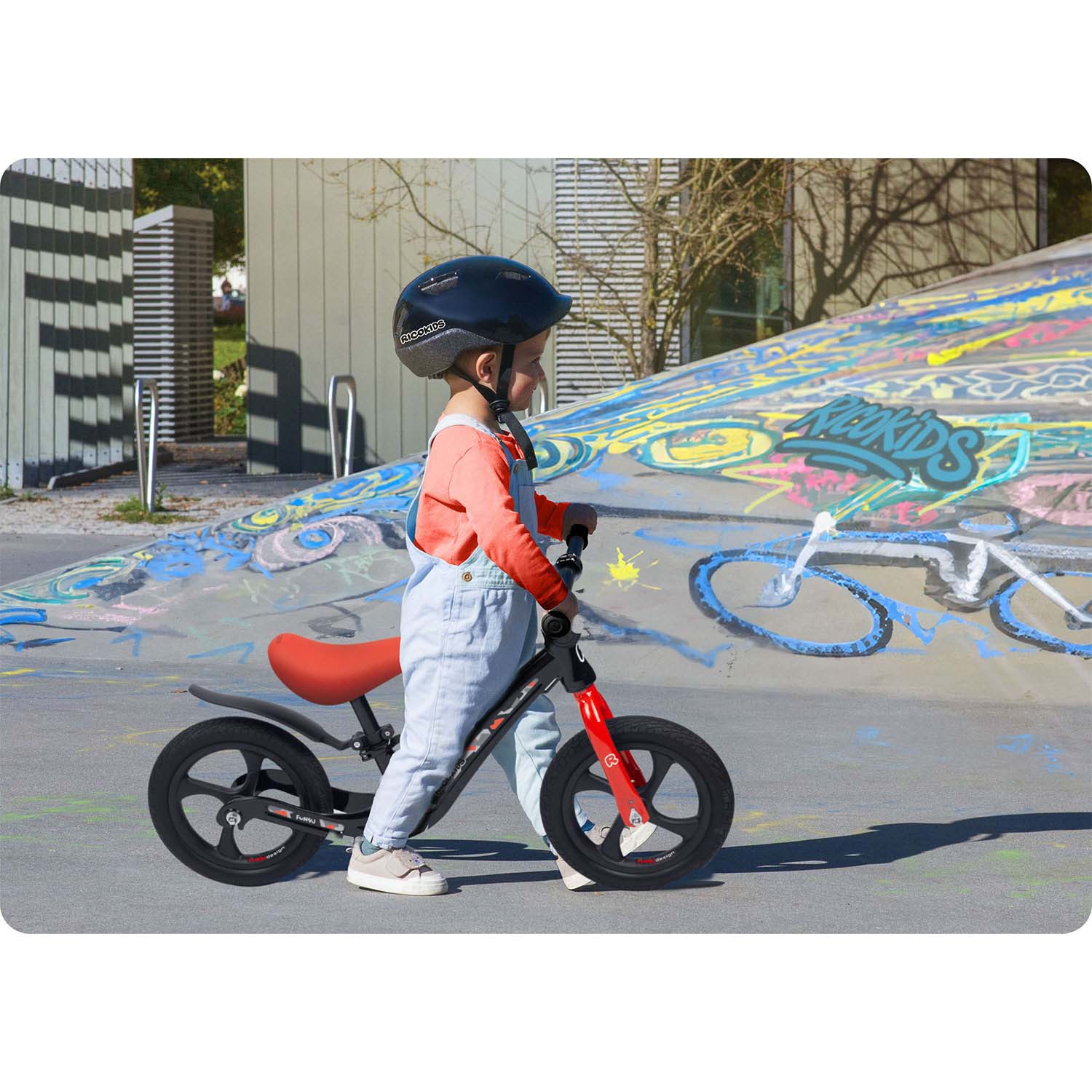 Dziecko w kombinezonie i kasku stoi obok czarno-czerwonego rowerka biegowego na utwardzonym terenie z graffiti.