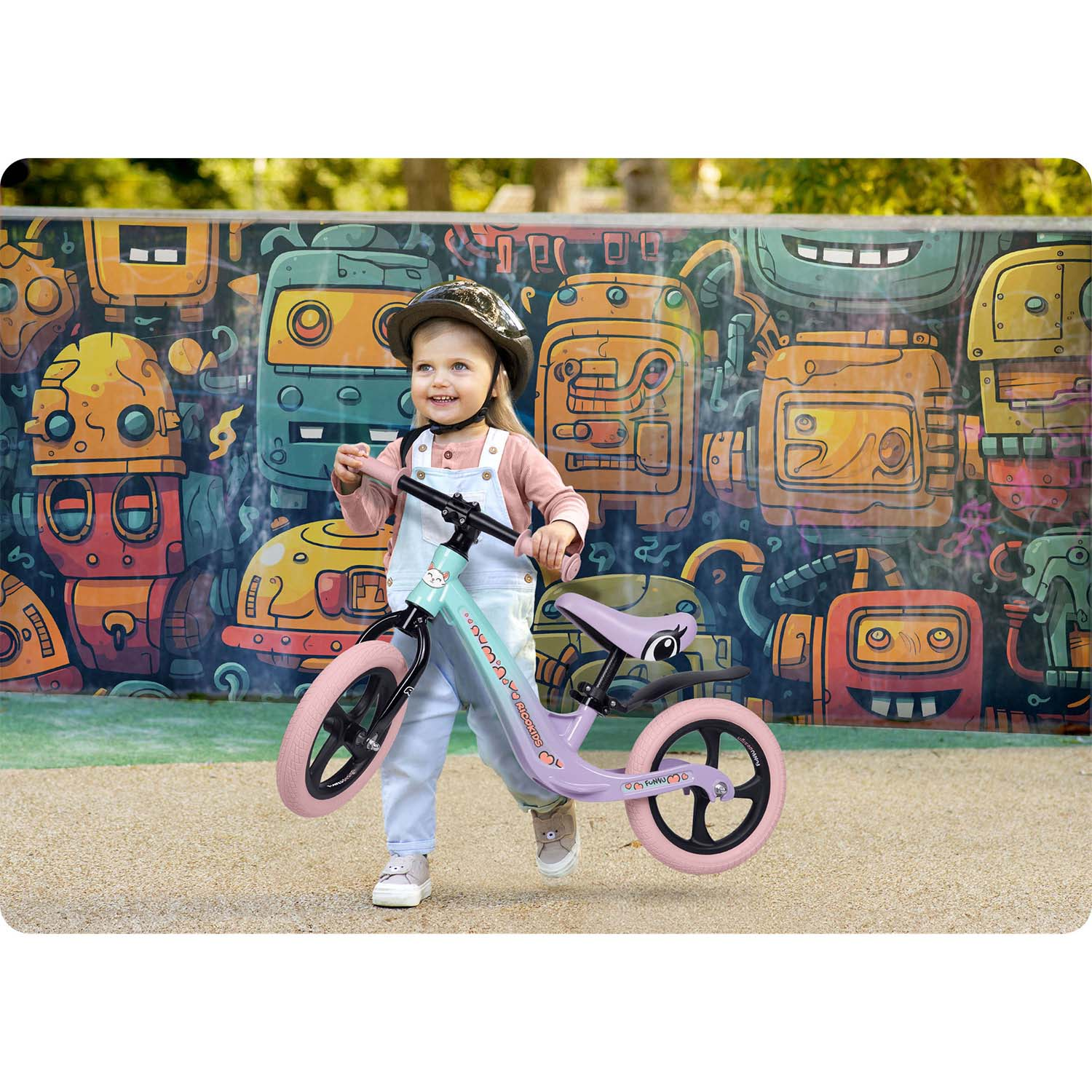 Mała dziewczynka w kombinezonie i kasku trzyma rowerek biegowy. Tło to kolorowy mural z postaciami przypominającymi roboty.