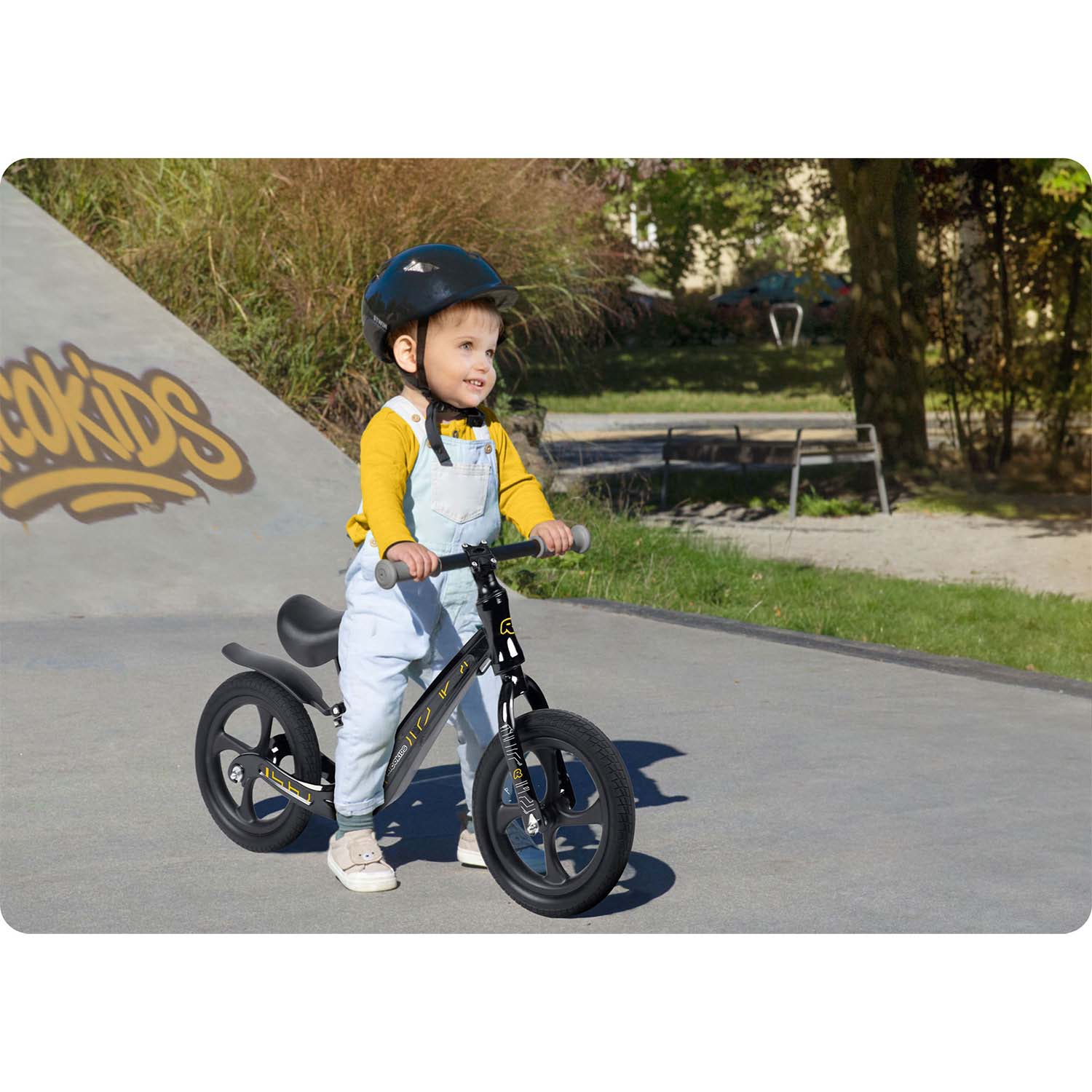 Dziecko na czarnym rowerze biegowym uśmiecha się. Ma na sobie kask i kombinezon. Tło obejmuje skatepark i drzewa.