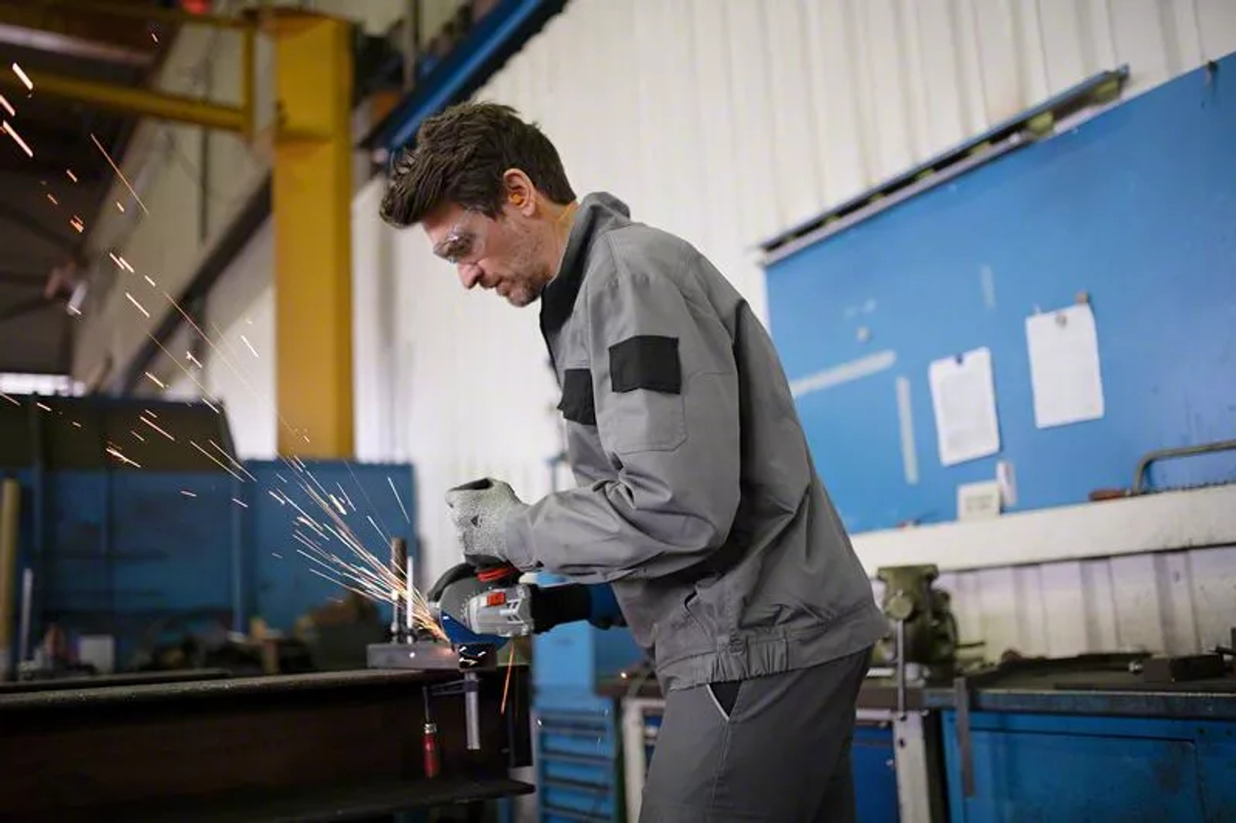 Robotnik szlifuje metal narzędziem, lecą iskry. Nosi okulary ochronne i szary kombinezon w warsztacie.