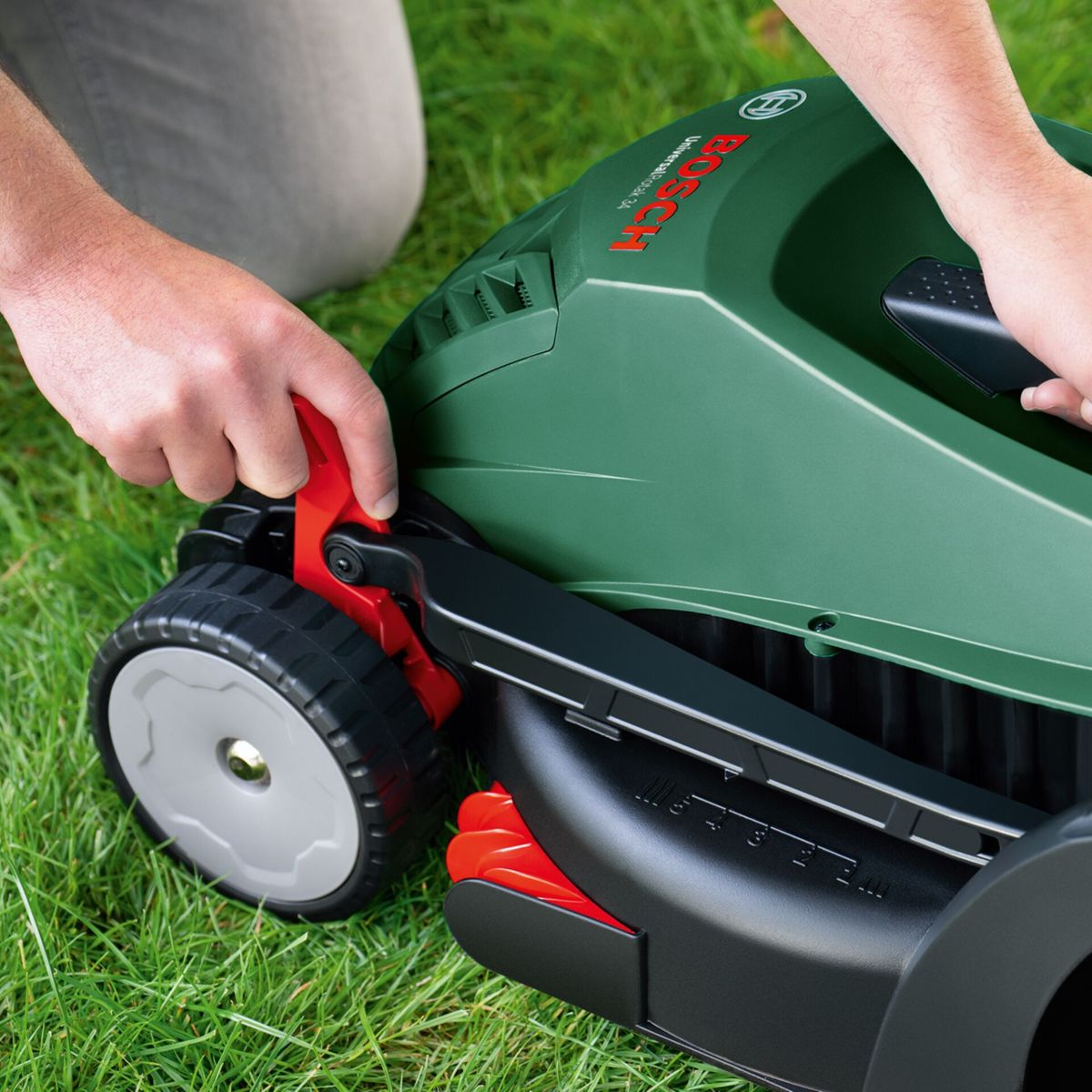 Eine Person bedient einen grünen Bosch-Rasenmäher auf Gras. Der Rasenmäher hat schwarze Räder und rote Akzente. Hände sind auf dem Mäher.