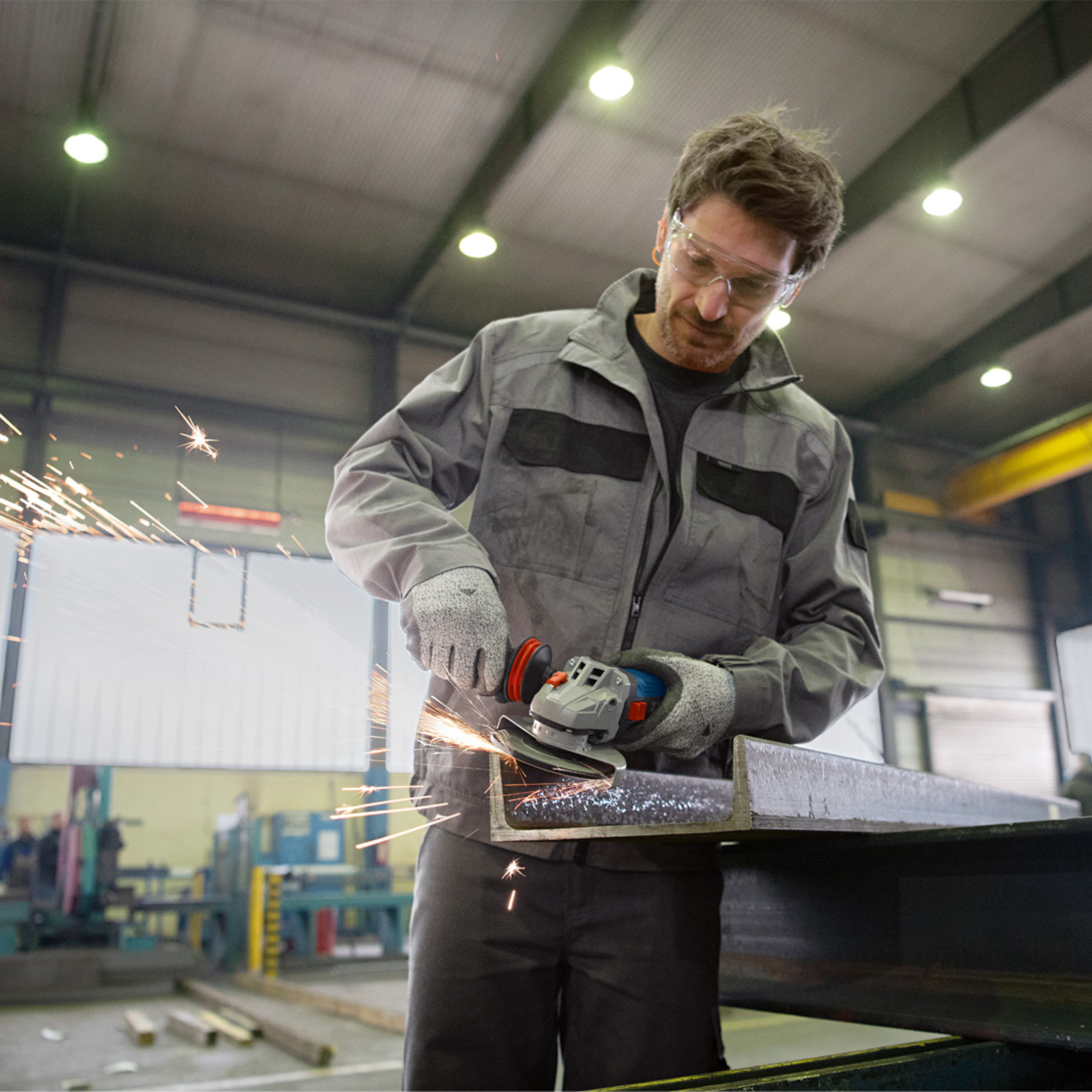 Mężczyzna szlifuje metal elektronarzędziem. Iskry lecą. Ma okulary ochronne, rękawice i szary kombinezon.