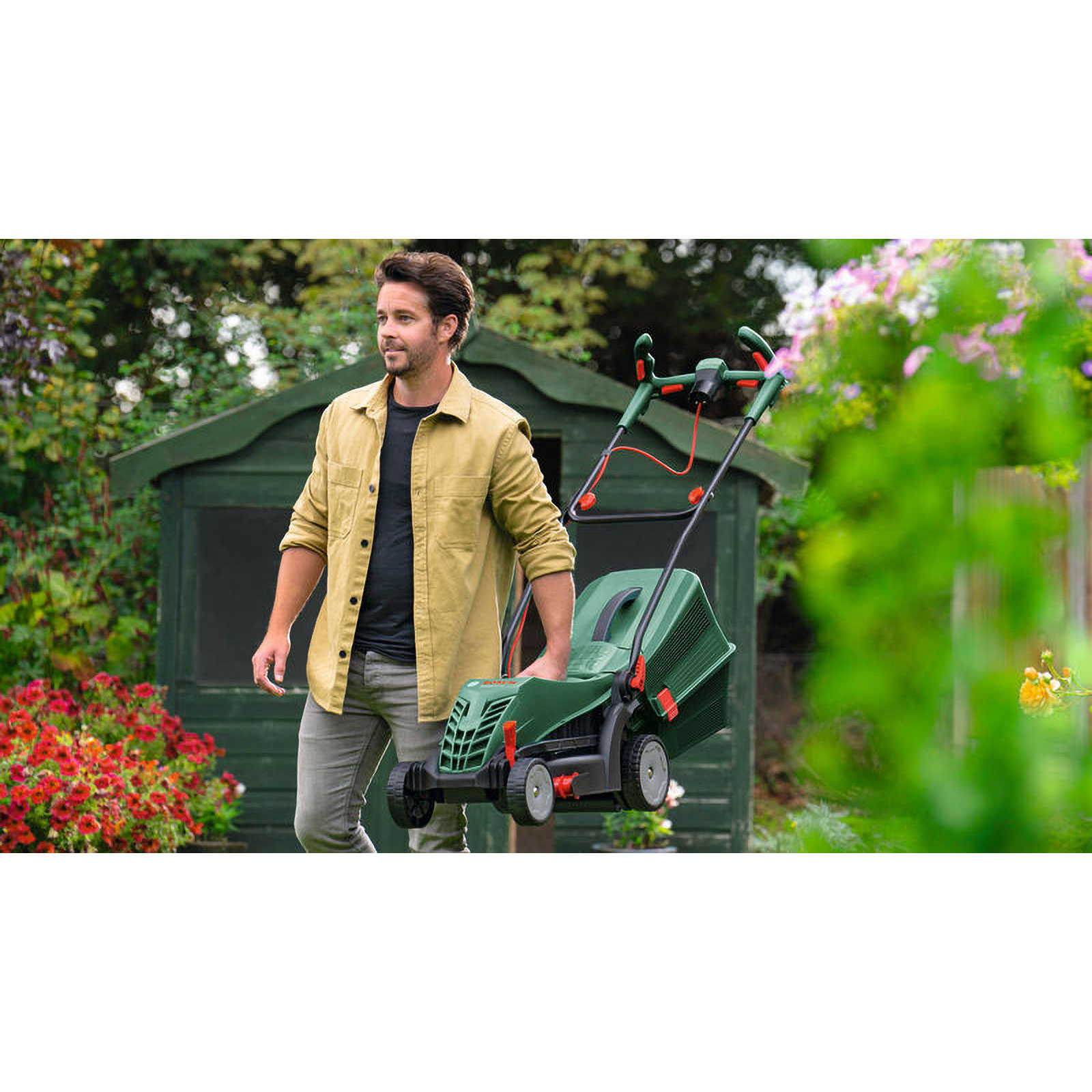 Ein Mann im Garten hält einen grünen Rasenmäher. Blumen und ein Schuppen sind im Hintergrund.