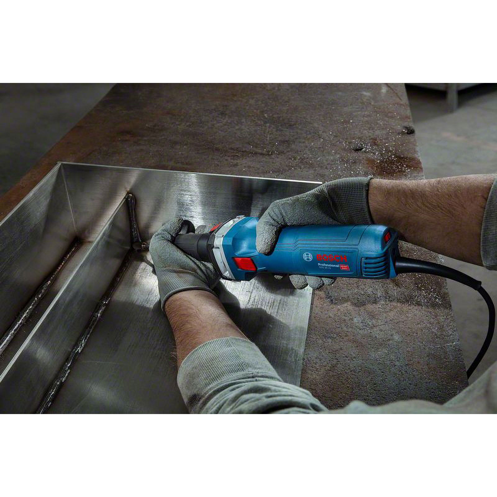 Bir işçi, eldiven takarak, metal bir yapı üzerinde mavi bir Bosch taşlama makinesi kullanıyor. Sahne bir atölyede geçiyor.