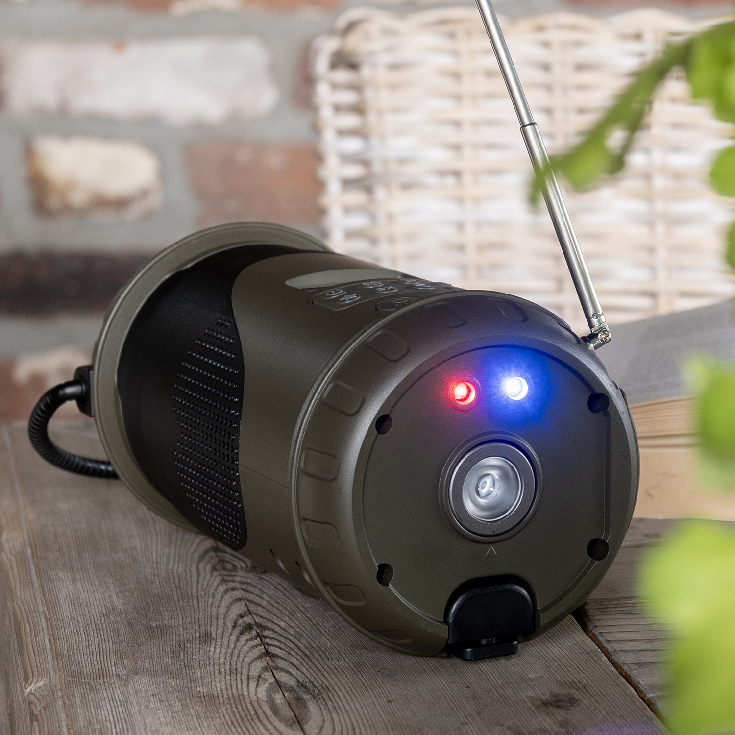 A dark green cylindrical device with red and blue lights sits on wood. It has an antenna and a speaker. A brick wall is in the background.