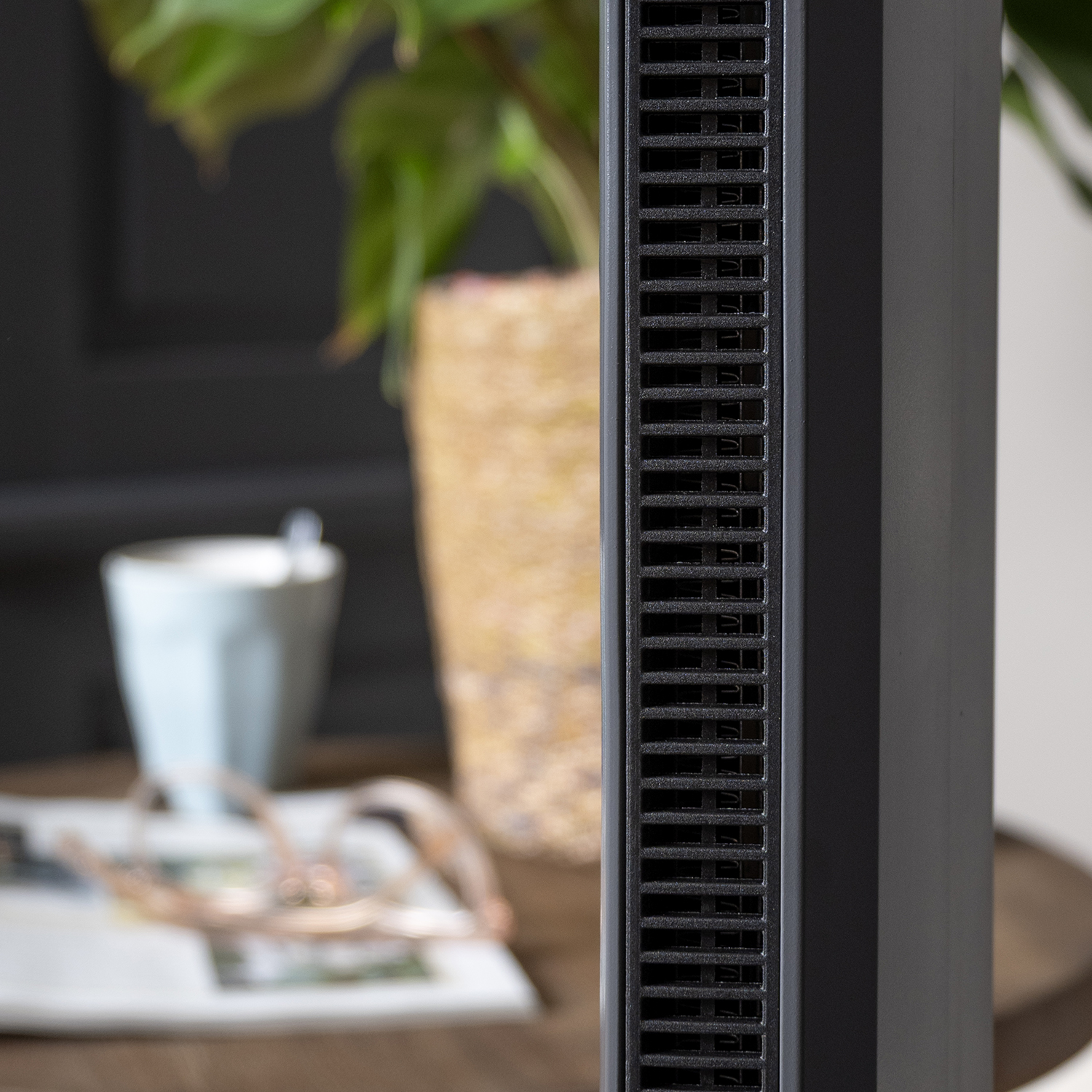 Close-up van een donkergrijs, rechthoekig apparaat met ventilatiesleuven. Vage achtergrond toont een mok, vaas en plant.