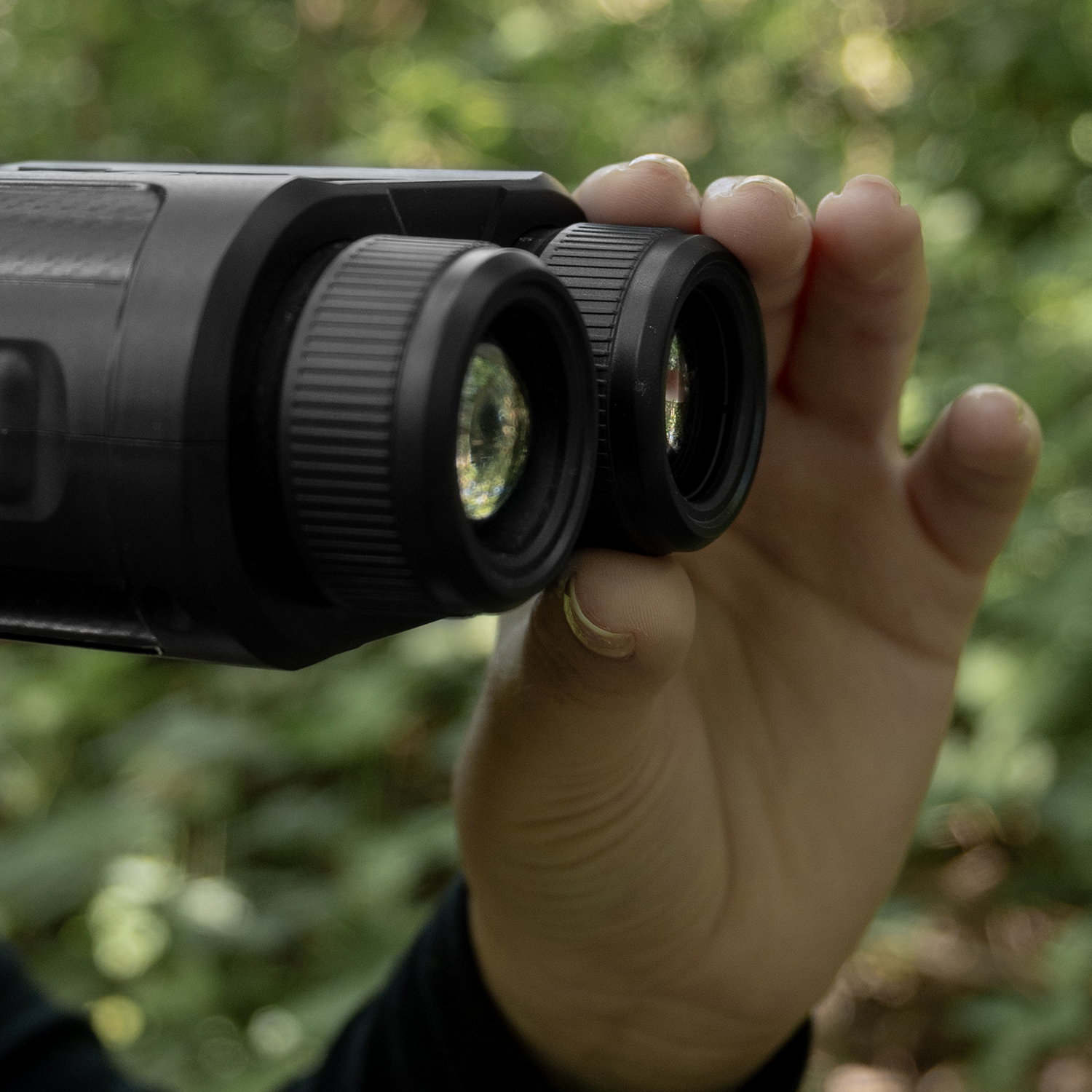 A person holds black binoculars, focused on lenses. The background shows blurred green foliage, suggesting an outdoor setting.