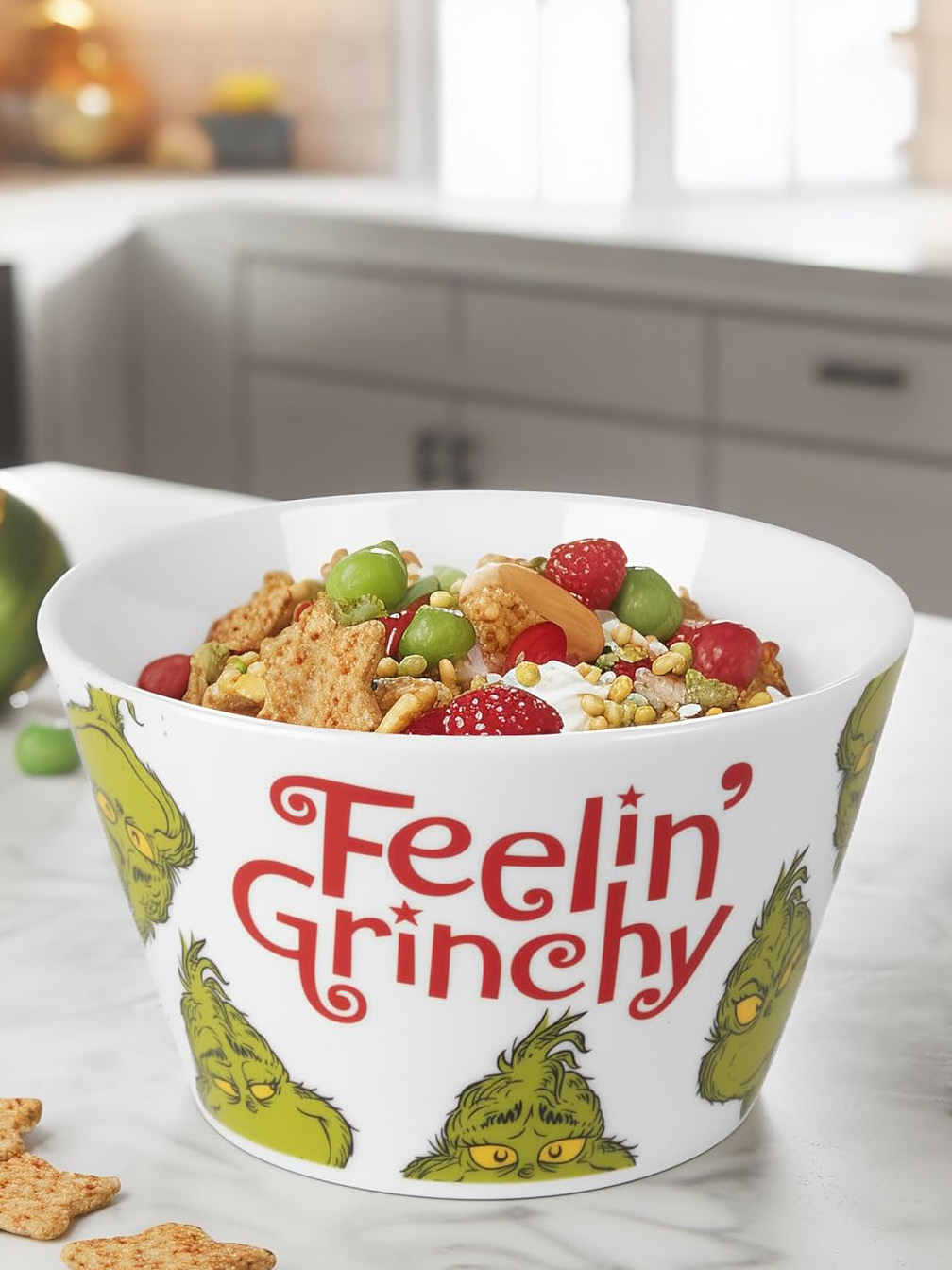 Eine weiße Schüssel mit dem Text "Feelin' Grinchy" und Grinch-Gesichtern, gefüllt mit Müsli und Obst, steht auf einem weißen Tisch.