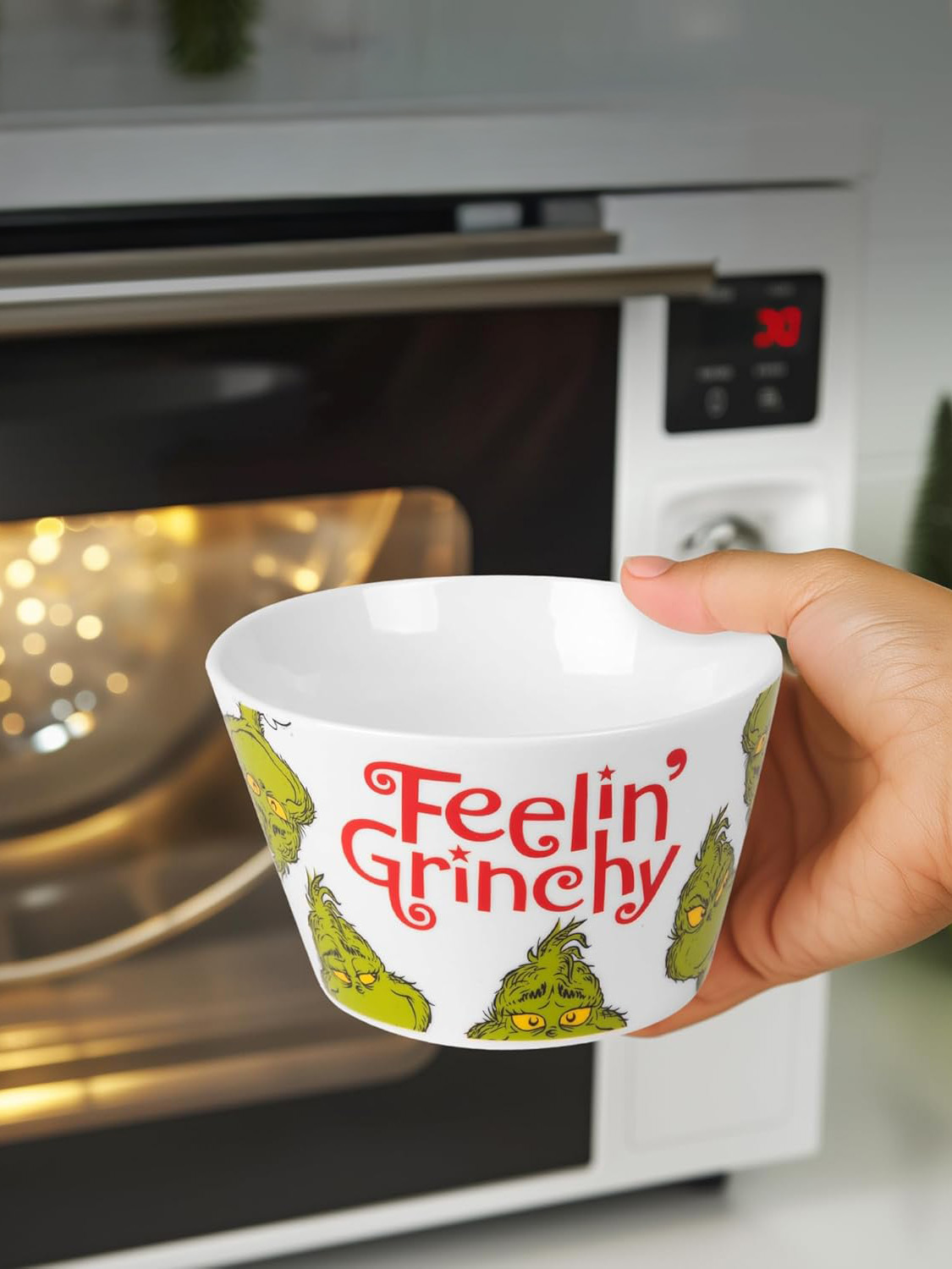 Eine Hand hält eine weiße Schüssel mit 'Feelin' Grinchy' und Grinch-Gesichtern. Im unscharfen Hintergrund ist ein Ofen zu sehen.