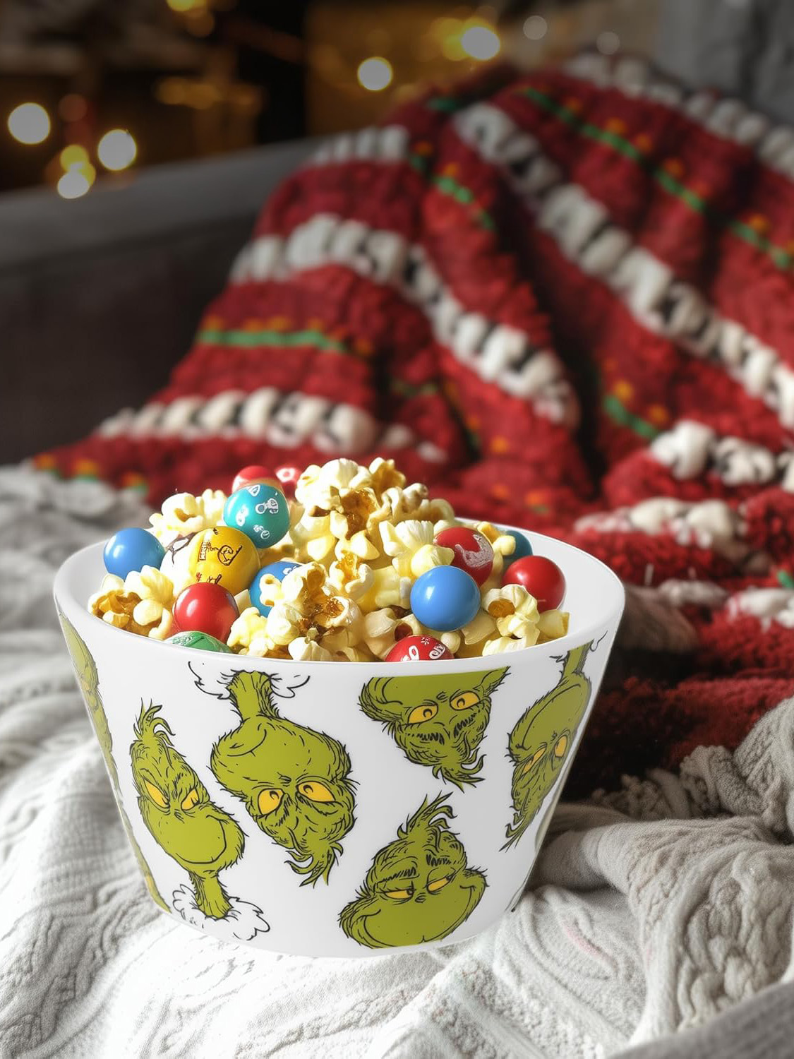 Eine Schüssel mit Grinch-Gesichtern, gefüllt mit Popcorn und bunten Süßigkeiten. Im Hintergrund befindet sich eine rot-weiß karierte Decke.