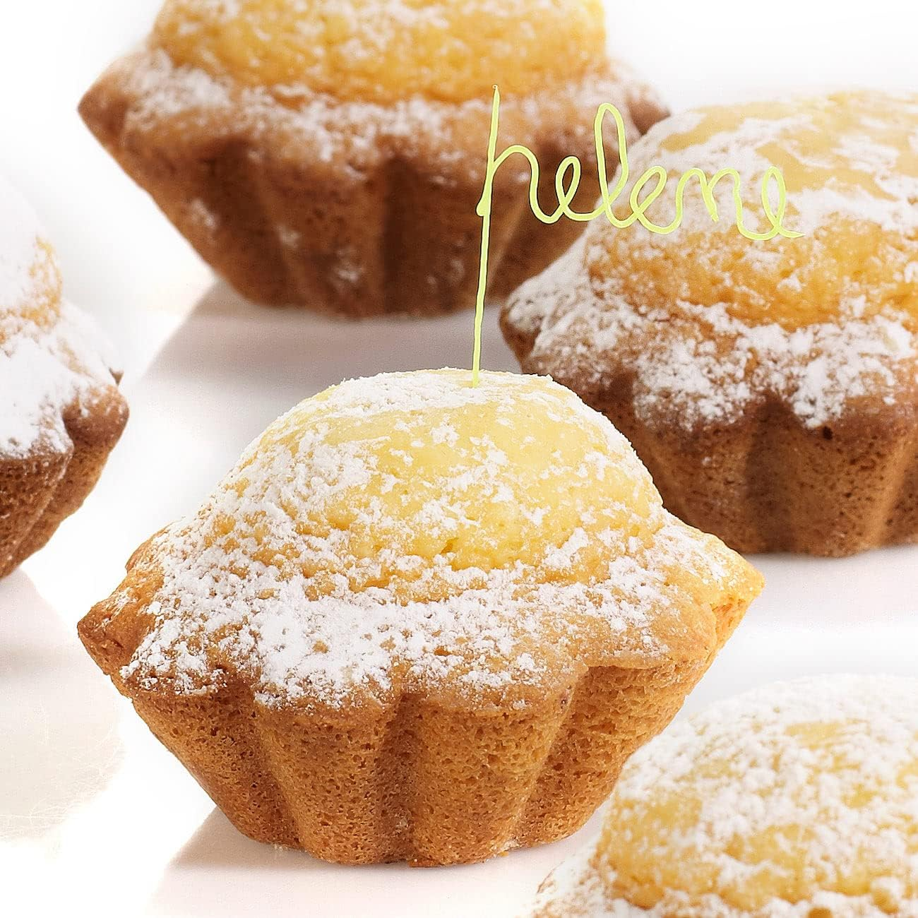 Primer plano de muffins espolvoreados con azúcar en polvo. Uno tiene un letrero de 'Helene'. El fondo es blanco.