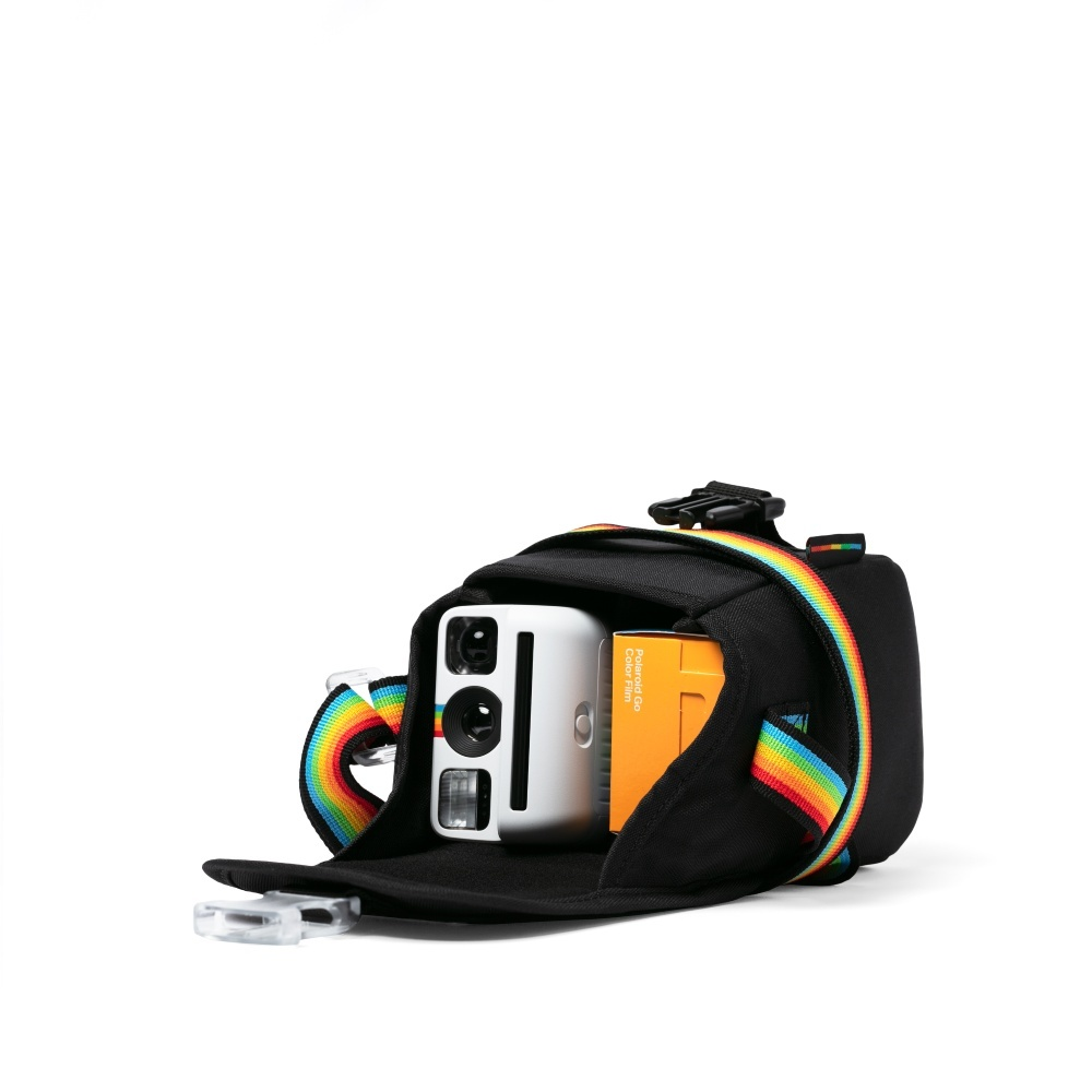 Eine schwarze Kameratasche mit Regenbogenriemen. Im Inneren sind eine weiße Polaroid-Kamera und eine orangefarbene Filmbox zu sehen.