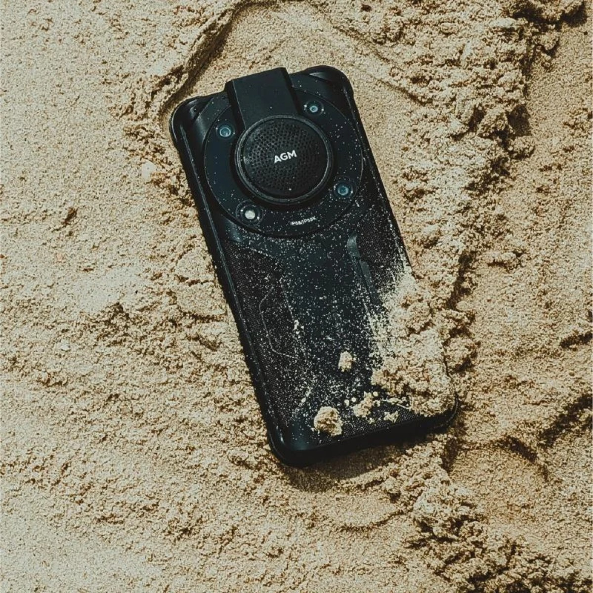 Uno smartphone nero con il logo 'AGM' giace sulla sabbia. La sabbia copre parzialmente il telefono. Lo sfondo è sabbioso.
