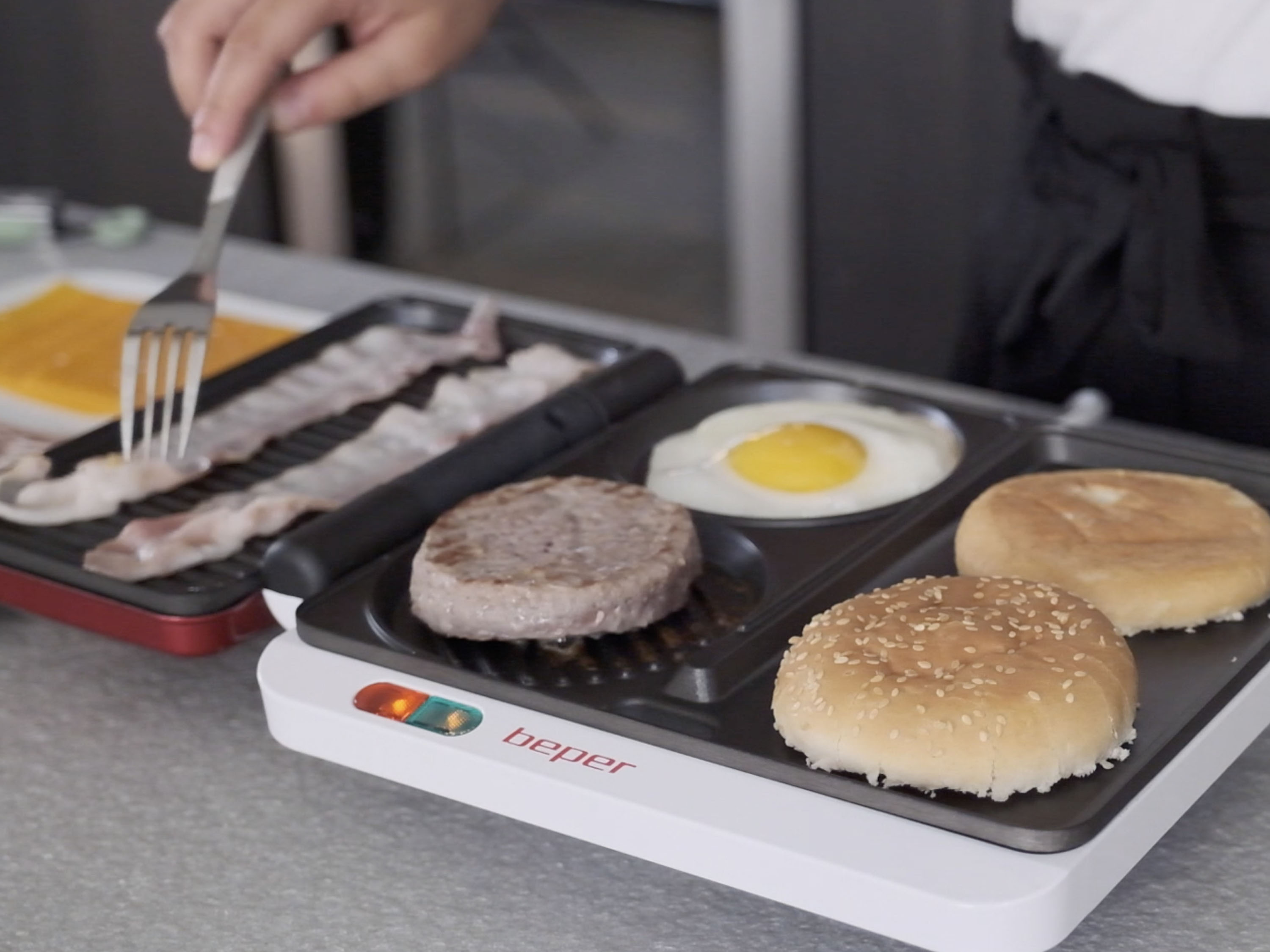 Una persona cucina un hamburger. Include pancetta, polpetta, uovo, panini su una piastra. Si usa una forchetta.