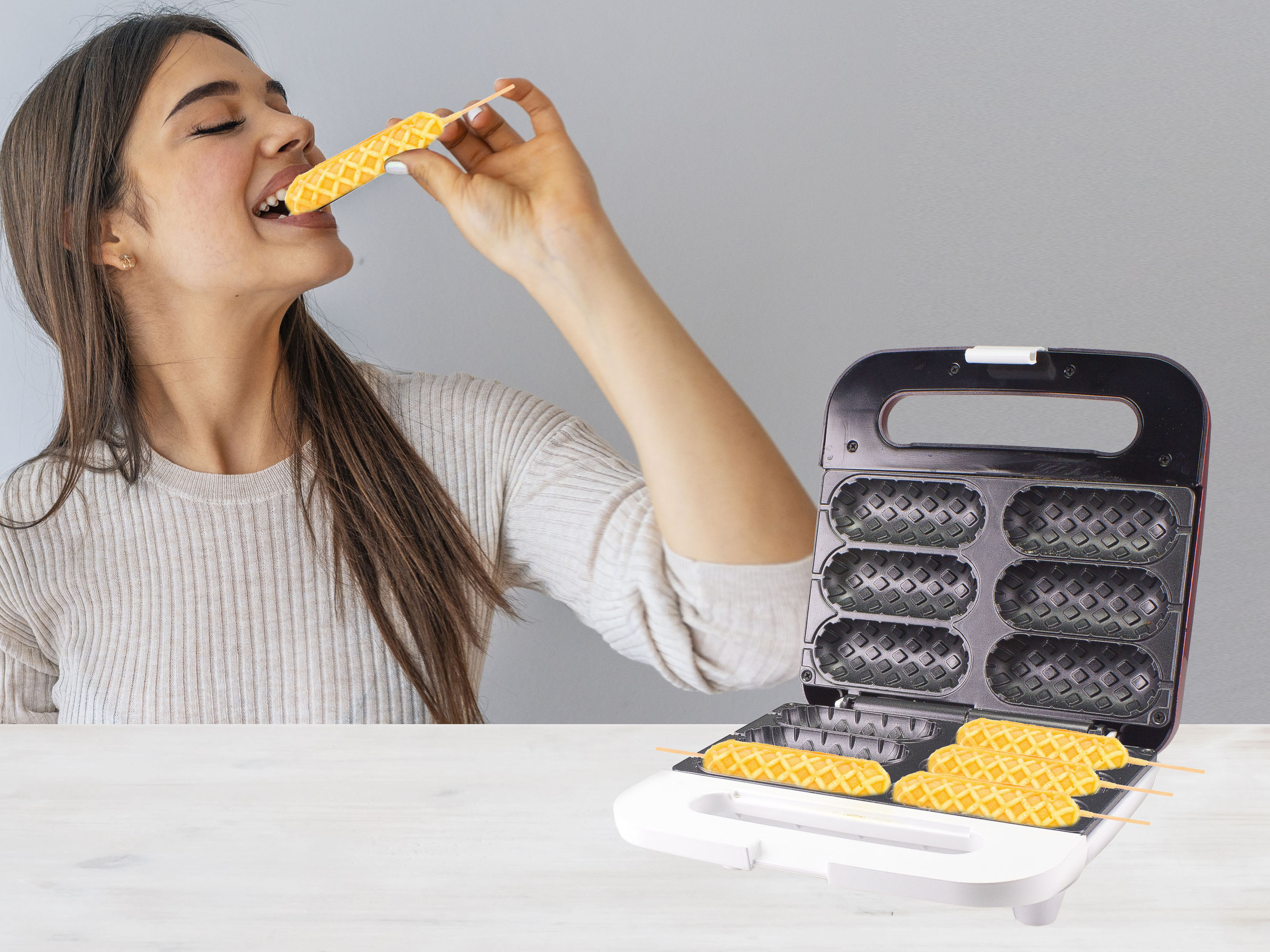 Eine Frau isst einen Waffelstab aus einem Waffeleisen. Der Maker ist geöffnet und zeigt weitere Waffelstäbchen.