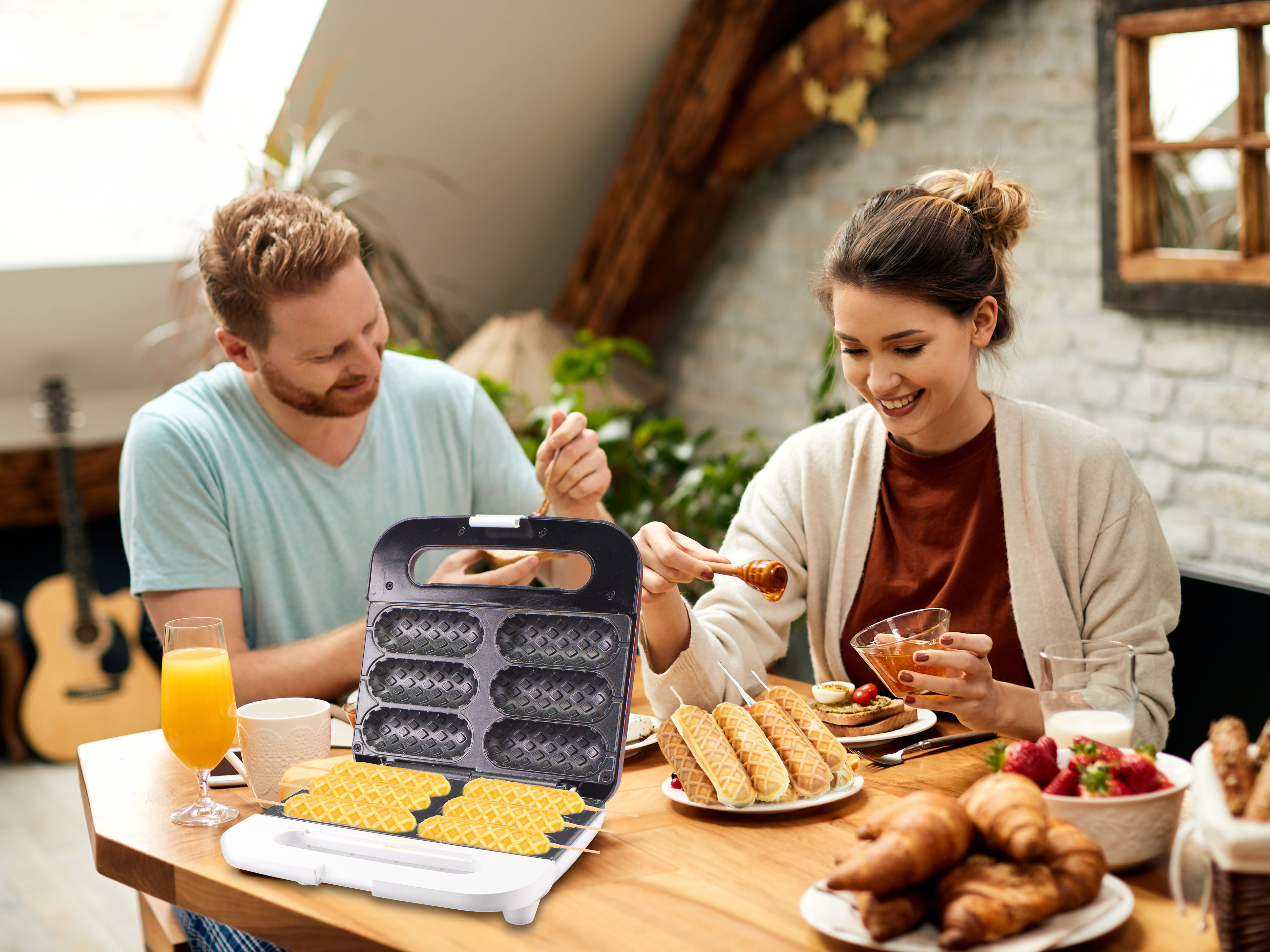 Ein Paar genießt ein Frühstück mit Waffeln, Honig, Croissants und Saft. Ein Waffeleisen ist geöffnet. Eine Gitarre ist im Hintergrund.