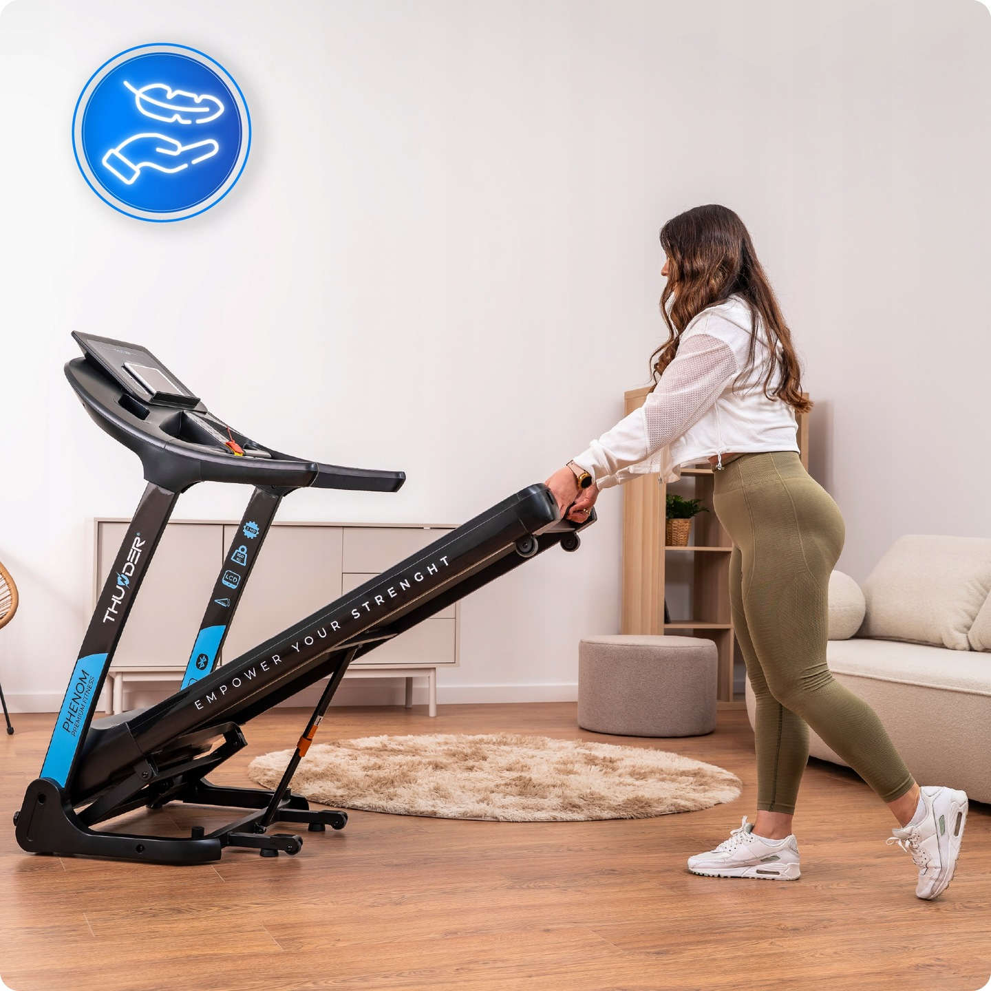 Kobieta w odzieży sportowej korzysta z bieżni. Bieżnia jest czarno-niebieska. Pokój ma drewniane podłogi i dywan.