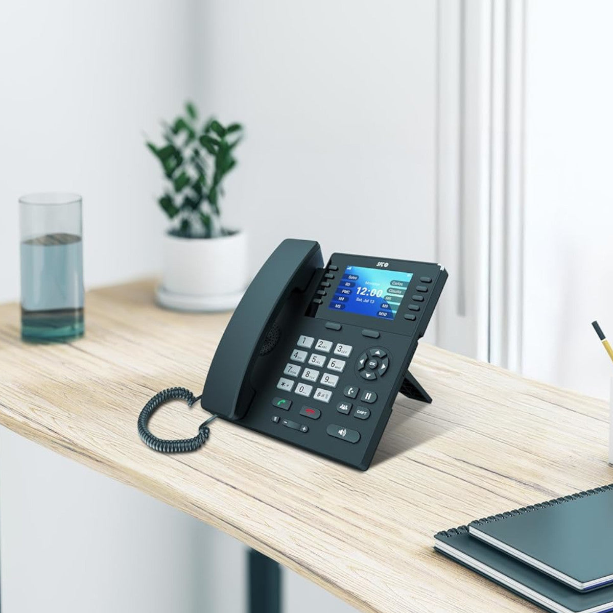 Un teléfono de oficina negro con pantalla en un escritorio de madera. Un vaso de agua y una planta en el fondo.