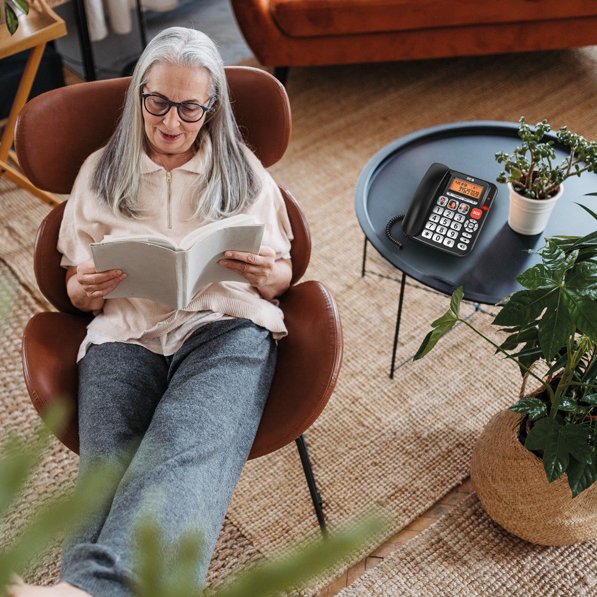 Una anciana con gafas lee un libro en una silla marrón. Un teléfono negro y plantas están en una mesa cercana.