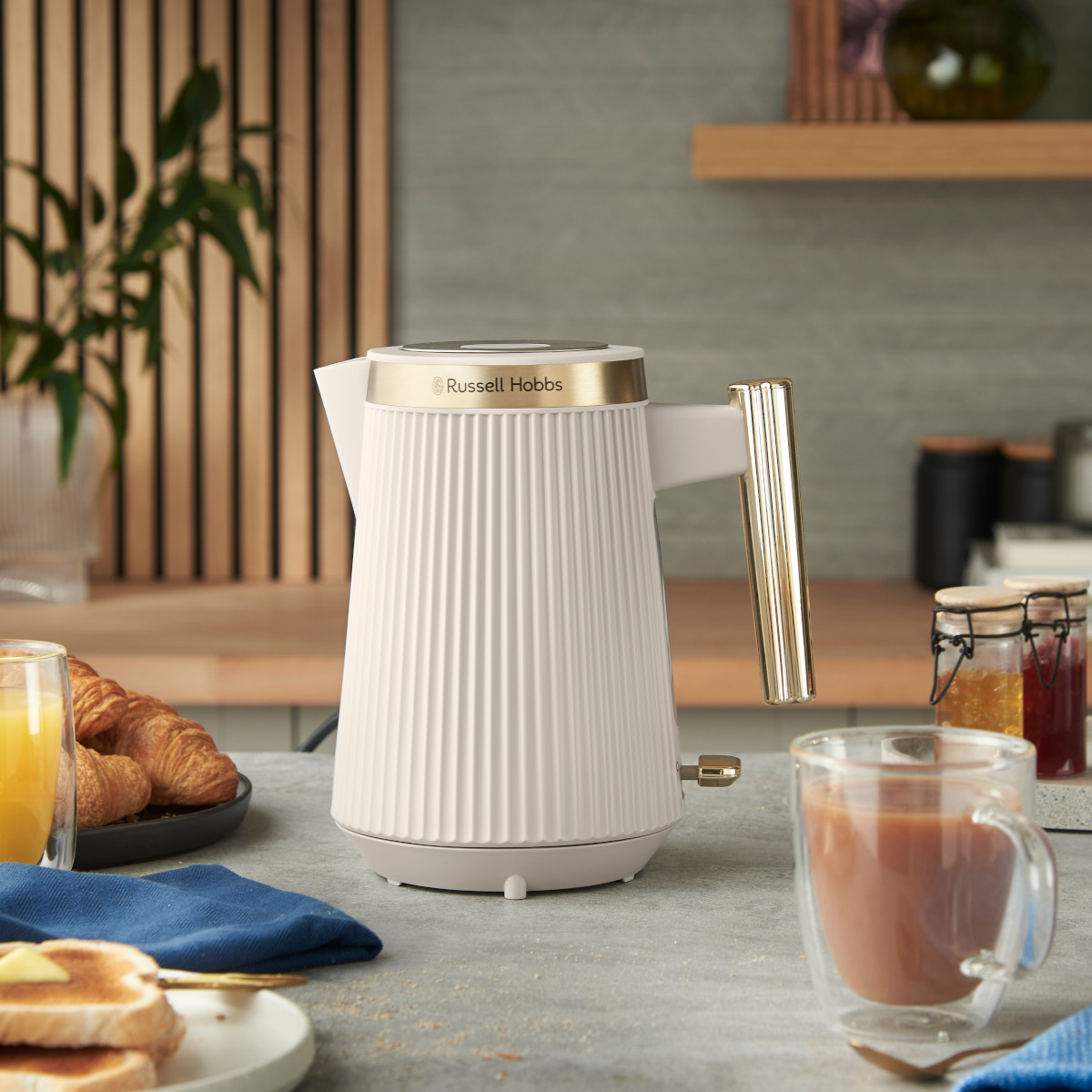 Biały czajnik Russell Hobbs na blacie ze śniadaniem i kawą. Tło ma panele drewniane i półki.