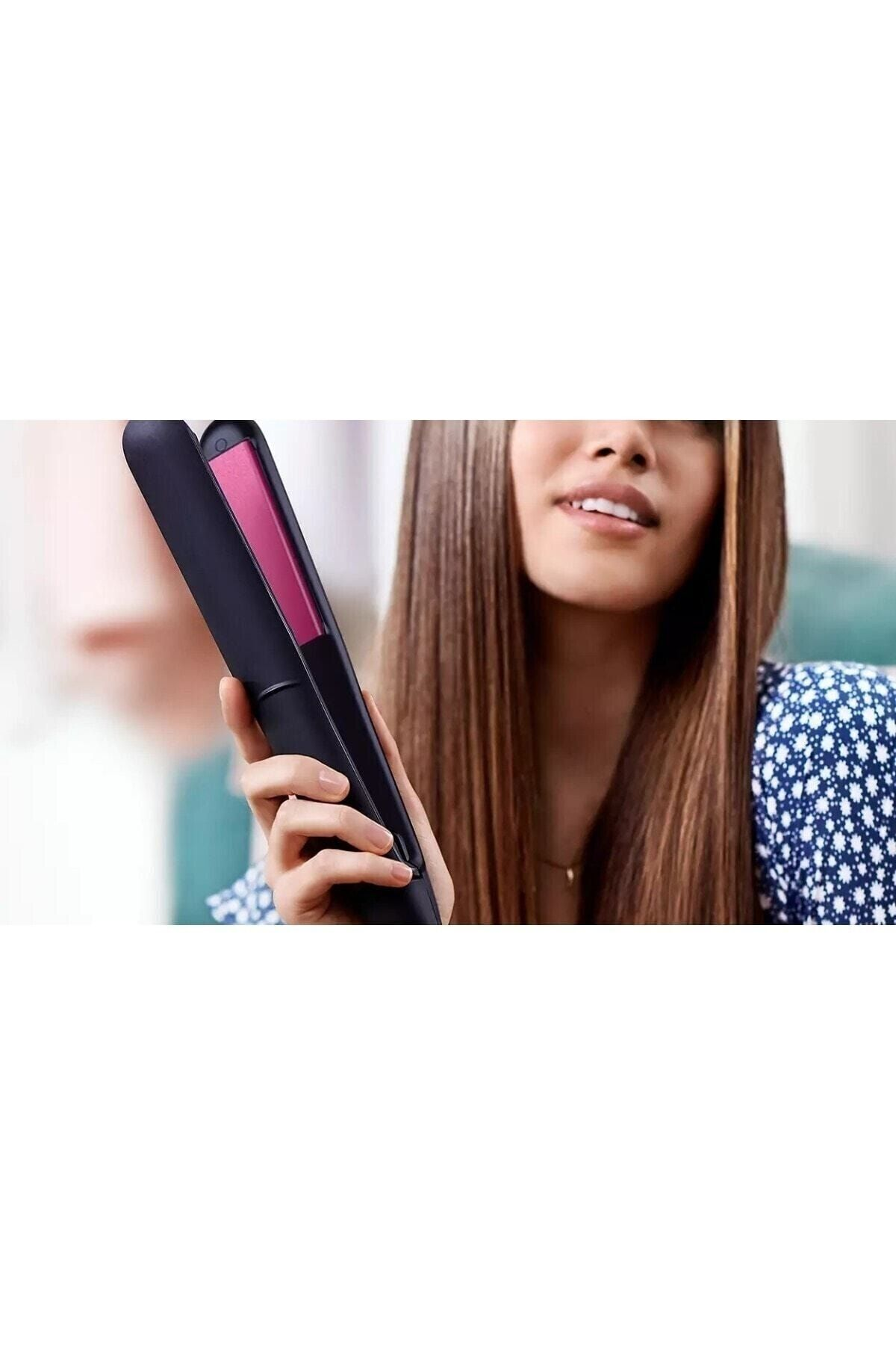 Kobieta trzyma czarną prostownicę do włosów z różową płytką. Ma na sobie niebieski top w kwiaty. Widoczne są długie brązowe włosy.