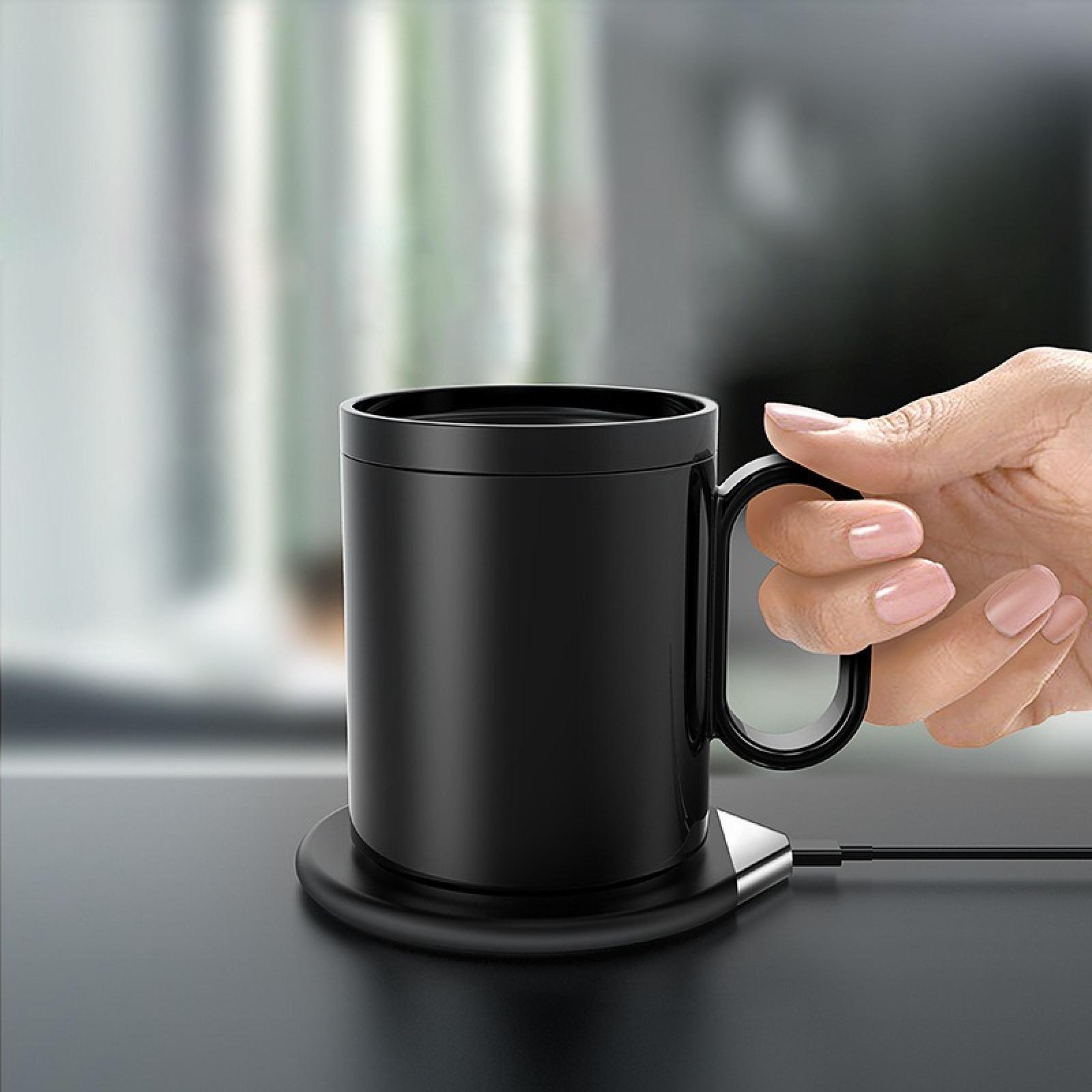 Eine schwarze Tasse auf einem Untersetzer wird von einer Hand mit rosa Nägeln gehalten. Ein Kabel ist eingesteckt.