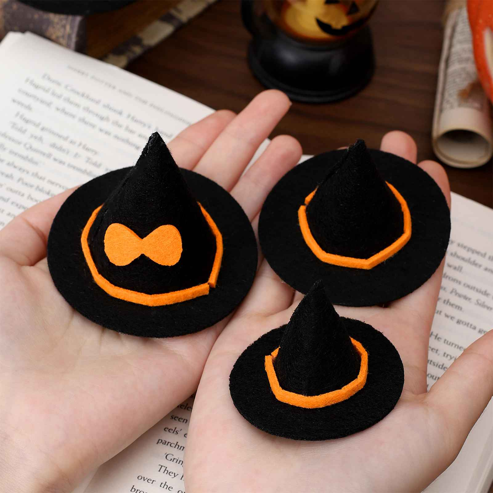 Hand hält drei Mini-Hexenhüte mit orangefarbenem Rand auf einer Buchseite. Ein Hut hat eine orangefarbene Schleife.