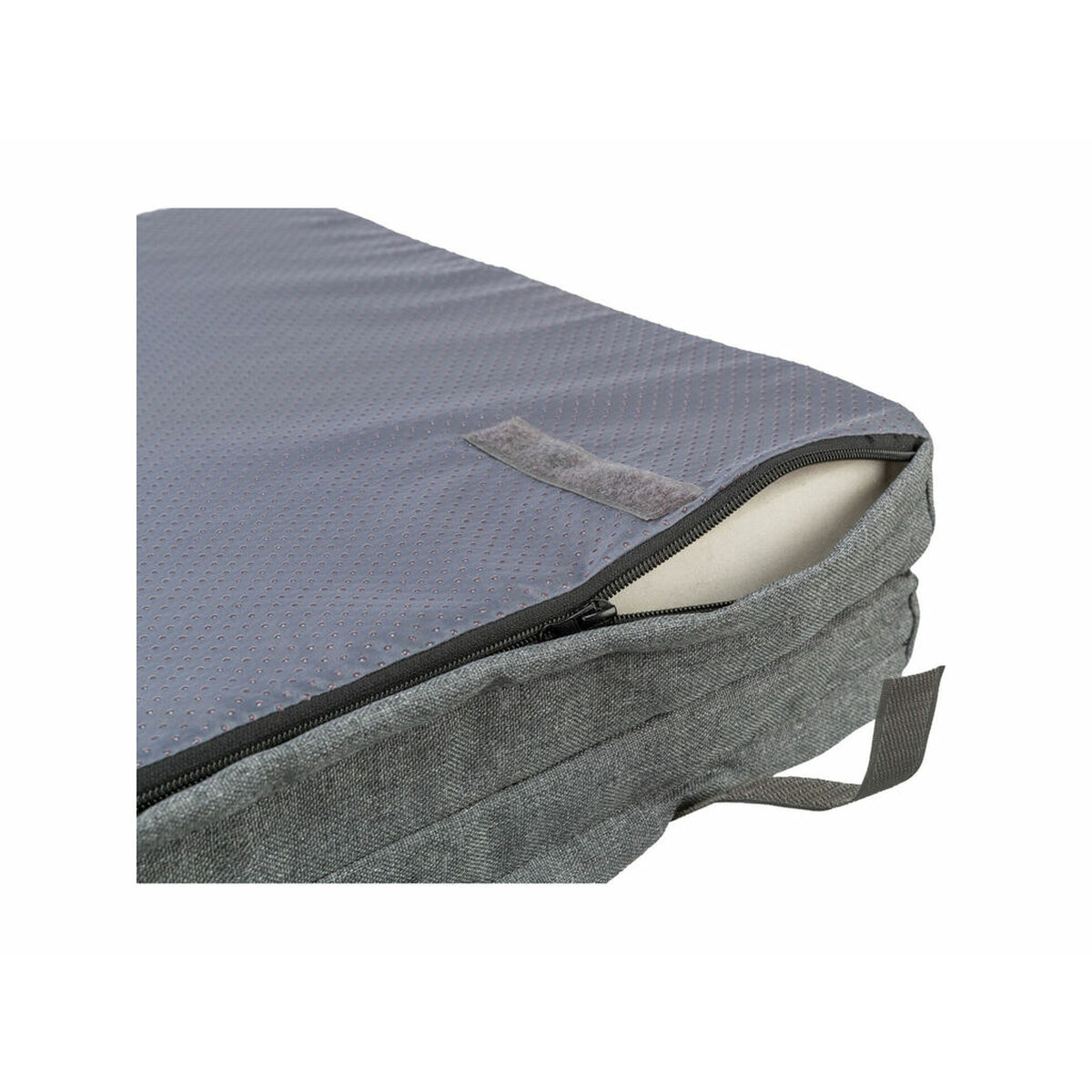Primer plano de una cama gris para mascotas. Una cremallera está parcialmente abierta, revelando espuma blanca. La parte superior tiene una superficie texturizada. Velcro visible.