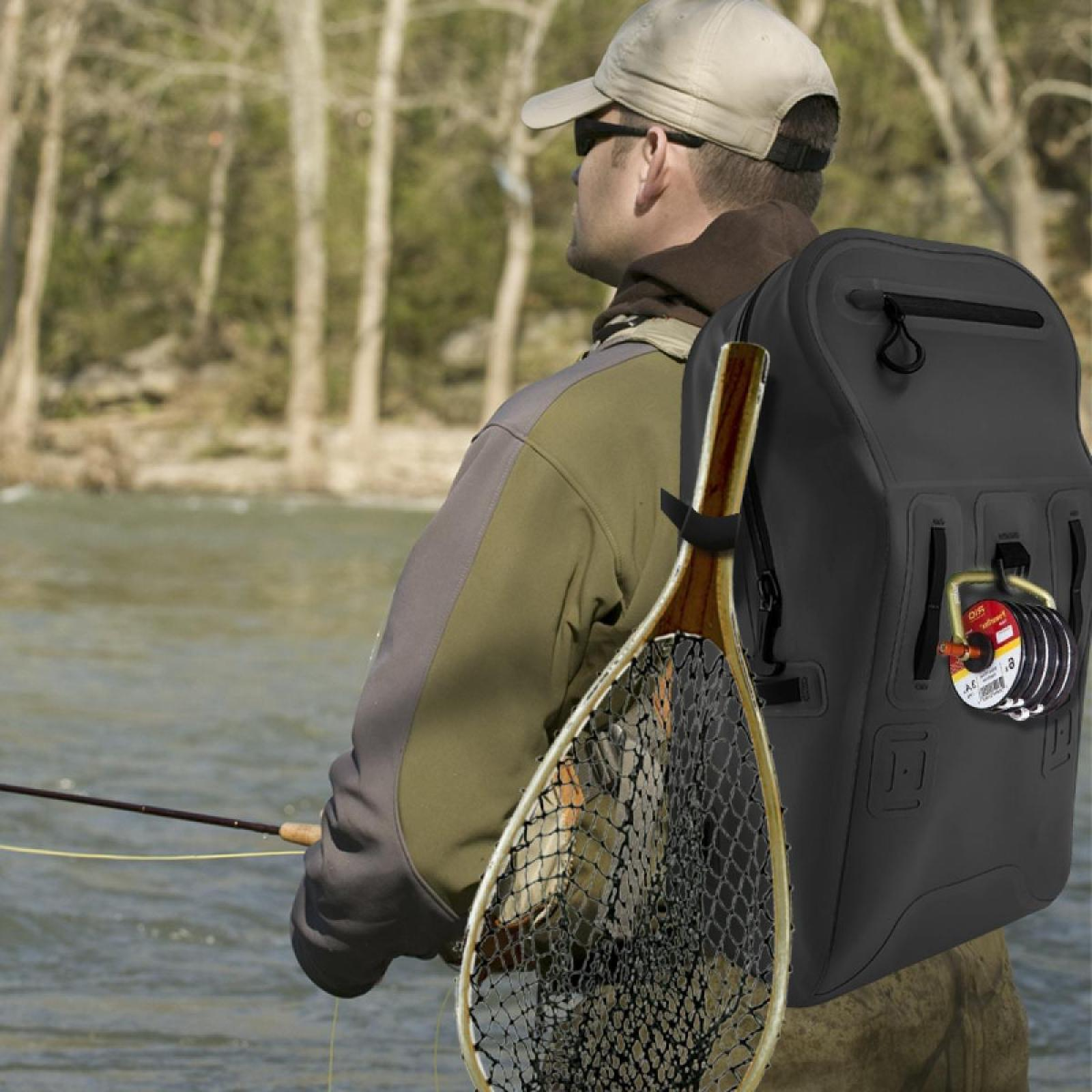 Ein Fischer steht in einem Fluss mit Rucksack und Fischernetz. Er hält eine Angel bereit zum Fischen.