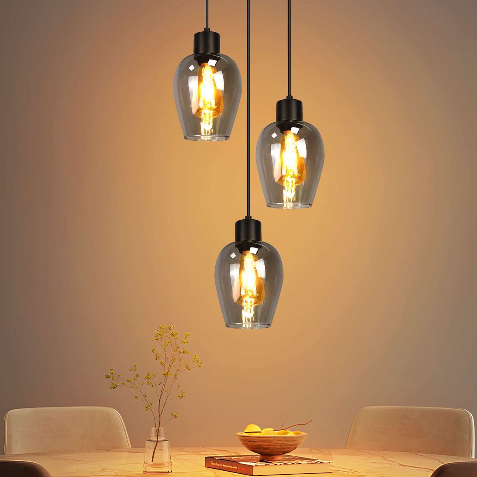 Tres lámparas colgantes de cristal cuelgan sobre una mesa con un jarrón y un cuenco de fruta. Iluminación cálida con fondo neutral.