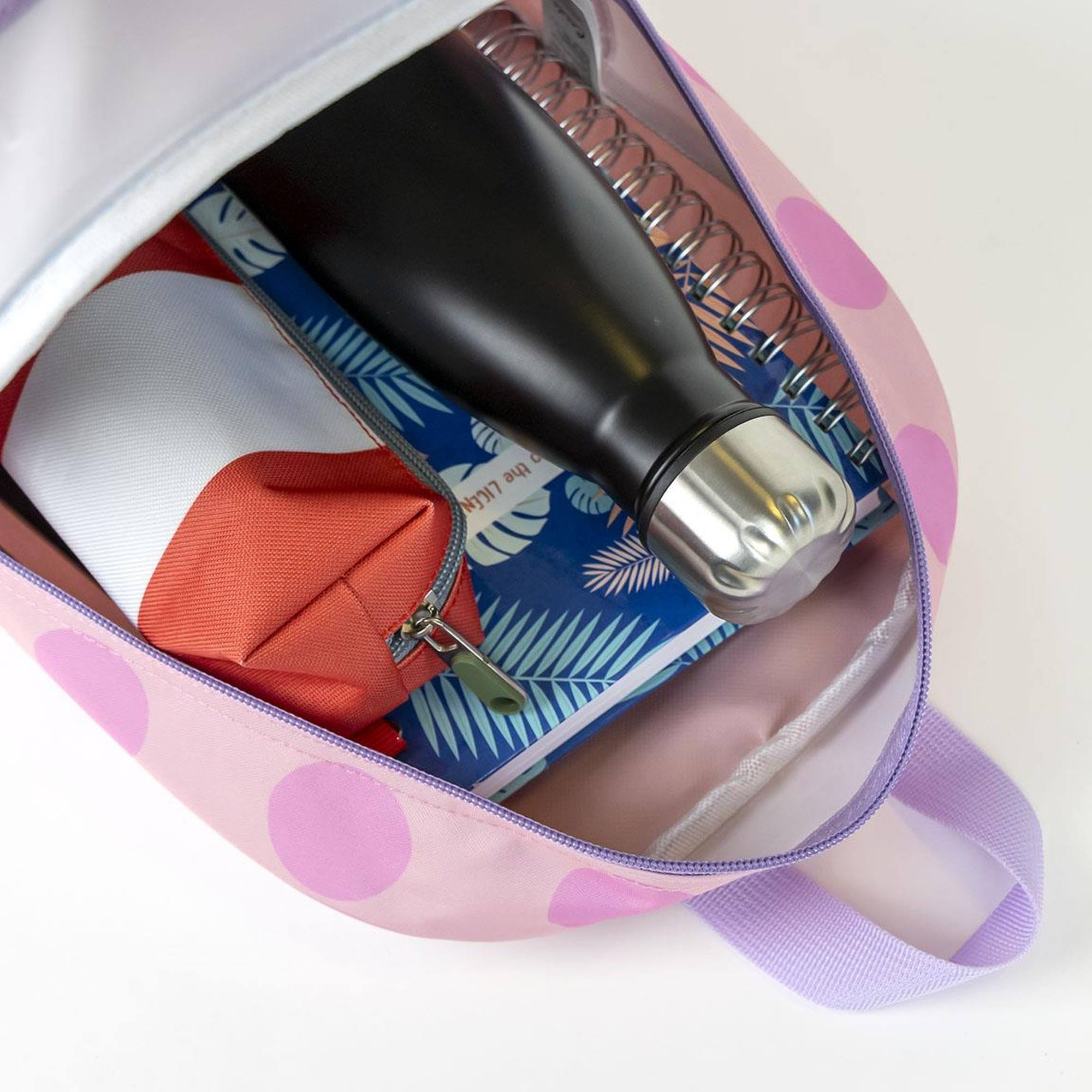 Offener rosa Rucksack mit Polka-Dots, der eine schwarze Flasche, ein Notizbuch und ein rot-weißes Federmäppchen zeigt.