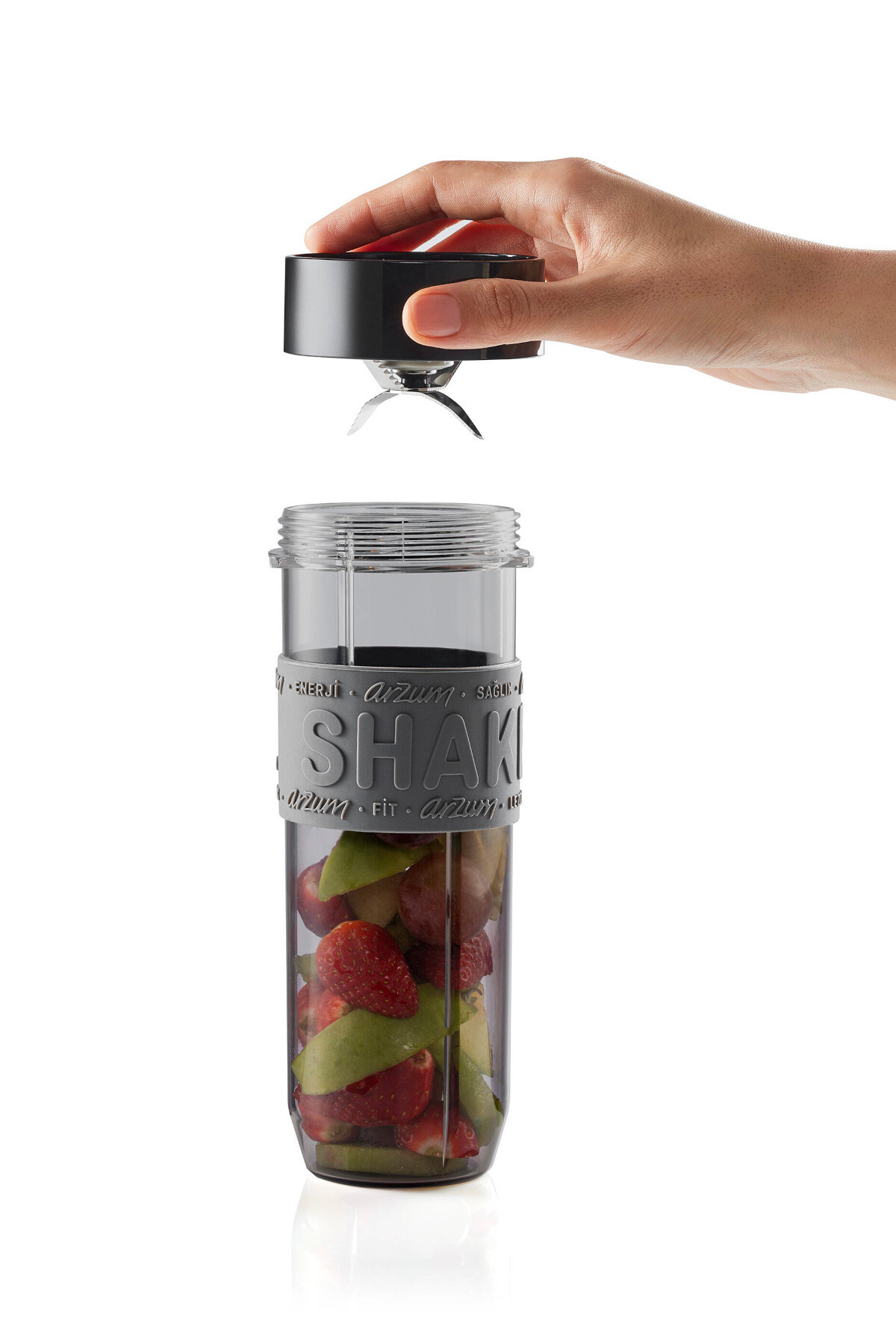Bir el, çilek ve yeşil meyvelerle dolu bir kabın üzerinde siyah bir blender kapağı tutuyor. Kapta 'SHAK' yazısı var.