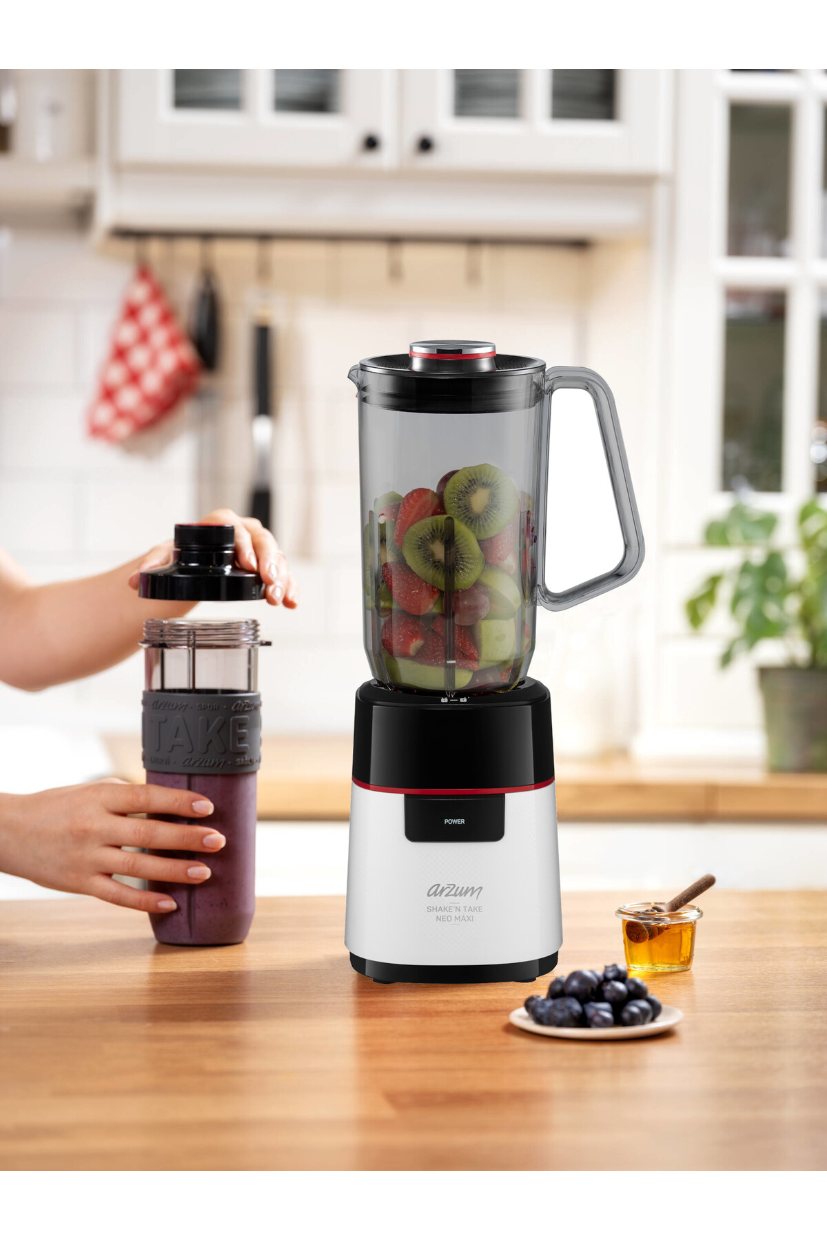 İçinde meyveler olan bir blender, bir bardak ve bir kişinin ellerinin yanında. Meyveler, bal ve yaban mersini ahşap bir masada.