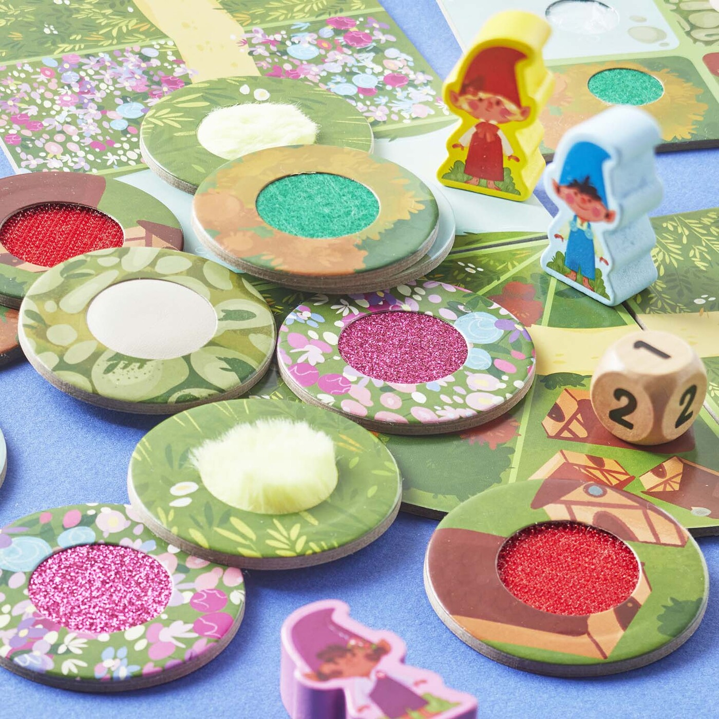 Brettspiel-Setup. Zwei Zwergfiguren, bunte Spielsteine, ein Würfel und ein Spielbrett auf blauem Untergrund. Blumen- und Glitzerakzente.