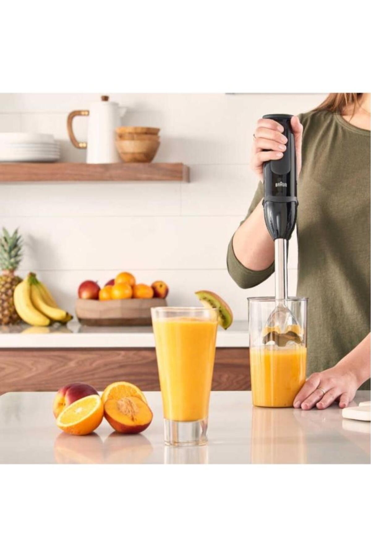 Bir kişi, bir daldırma blender ile suyu karıştırıyor. Tezgahta portakal, şeftali ve kivi gibi meyveler var. Bir bardak meyve suyu mevcut.