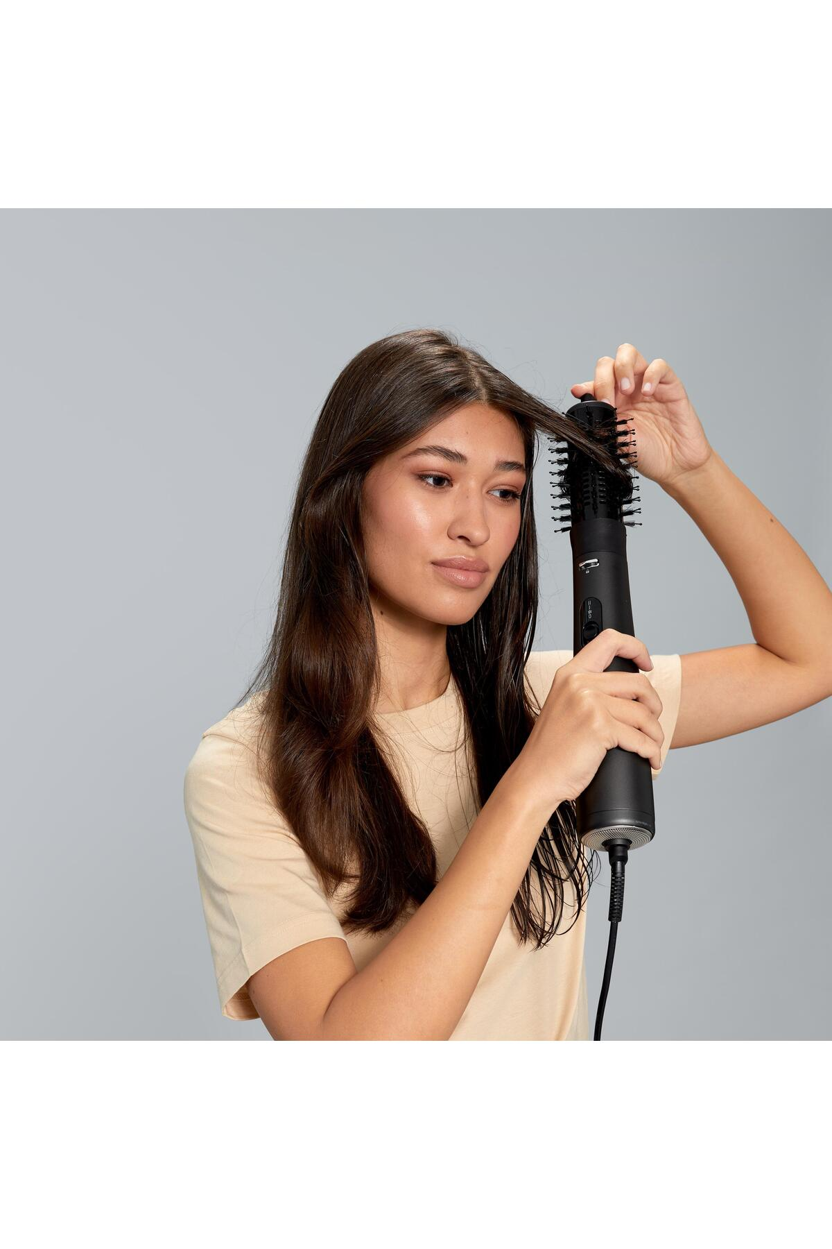 Una donna con lunghi capelli castani usa una spazzola nera per capelli. Indossa una maglietta beige chiaro su sfondo grigio.