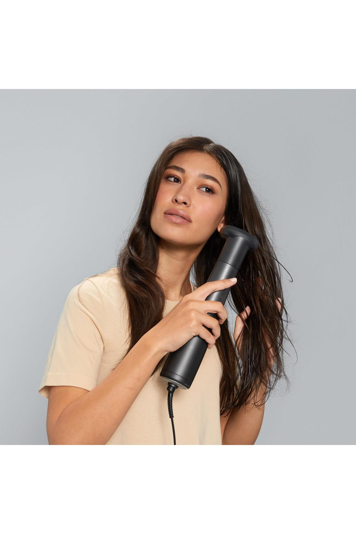 Una donna usa un asciugacapelli nero sui suoi capelli bagnati. Indossa una camicia beige. Lo sfondo è grigio chiaro.