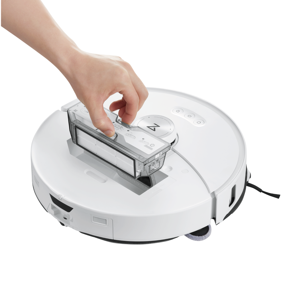 Ręka wyjmuje pojemnik na kurz z białego robota odkurzającego. Odkurzacz ma okrągły kształt. Pojemnik jest przezroczysty.