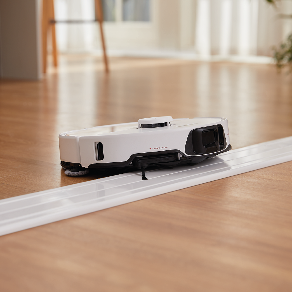 Biało-czarny robot odkurzający na drewnianej podłodze, w pobliżu białego toru. Tło z oknem i roślinami.