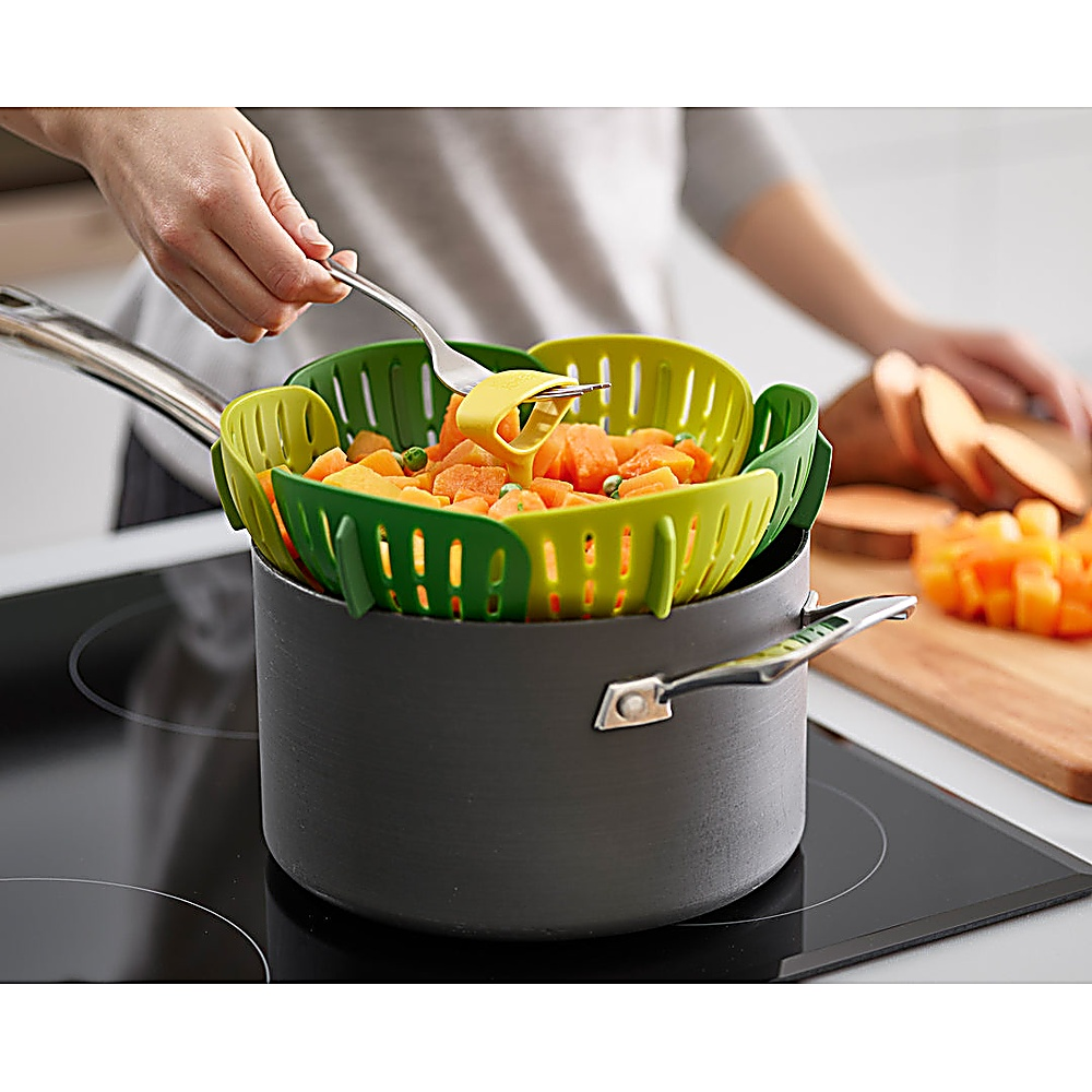 Een persoon gebruikt een vork om groenten uit een groen en geel stoommandje in een pot te halen. Zoete aardappelen staan op de achtergrond.