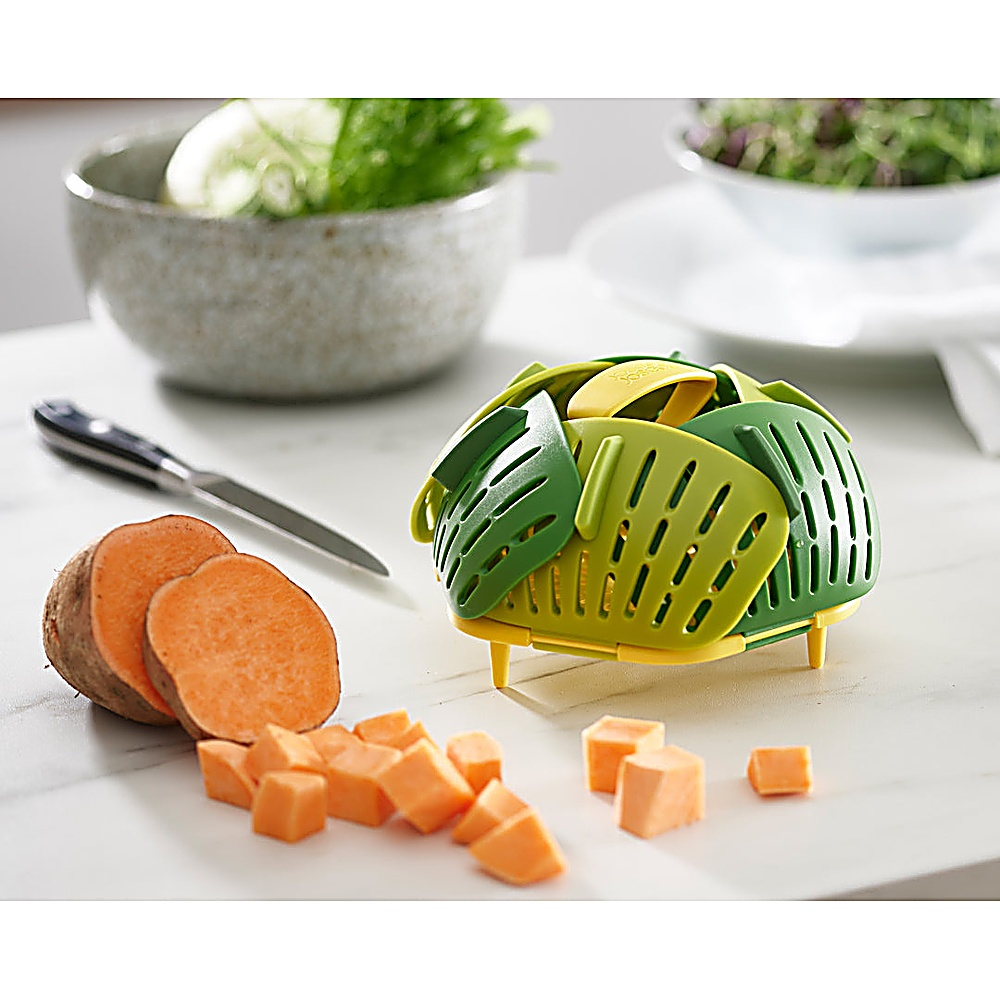 Een groene en gele groentestomer staat op een wit oppervlak. Zoete aardappelen zijn in de buurt gesneden. Schalen met groenten zijn op de achtergrond.