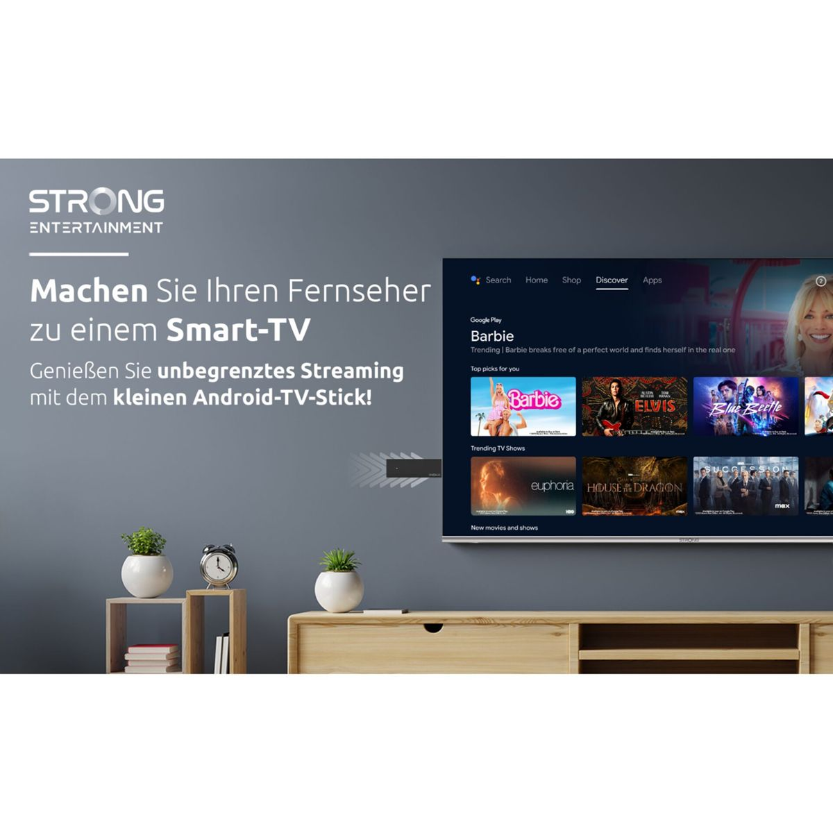 Pantalla de TV mostrando la película Barbie, conectada a un pequeño Android TV stick. Mueble de madera con plantas y reloj.