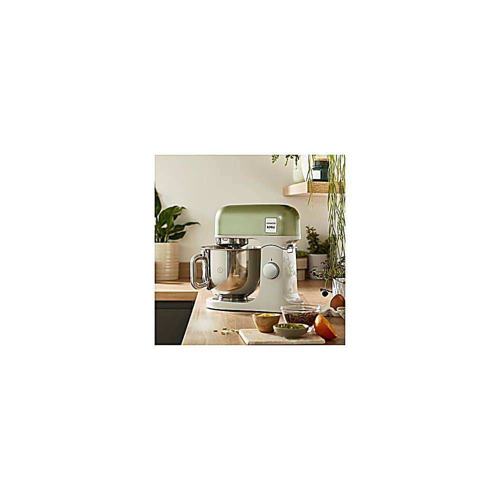 Ein weiß-grüner Küchenmixer steht auf einer Holztheke. Schüsseln und Pflanzen sind im Hintergrund. Eine Orange ist geschnitten.